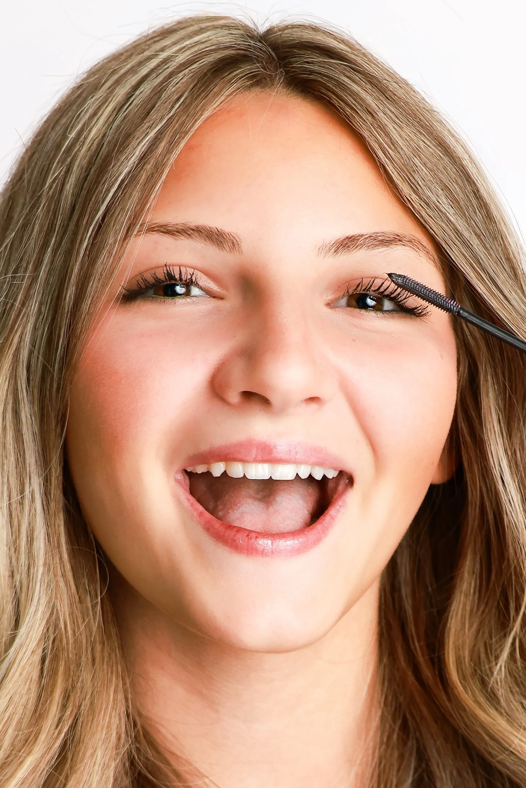 A teen model smiling with her mouth open while applying mascara to her right eyelash.