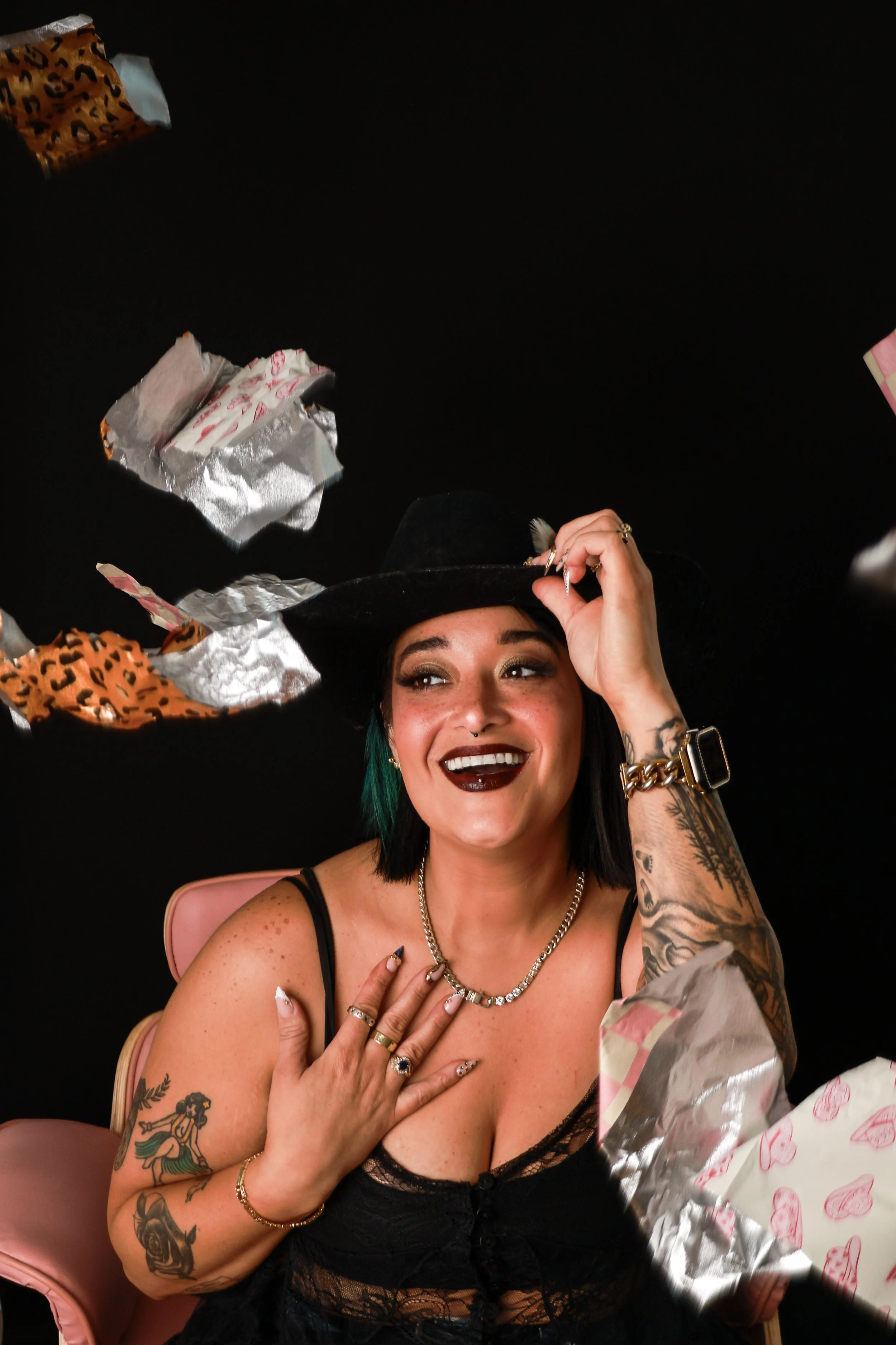 A woman with short black hair and tattoos on her arms, wearing a black lace top, gold jewelry, and a black hat, sitting in a pink chair, smiling and touching her chest, surrounded by wrapped gifts on a black background.