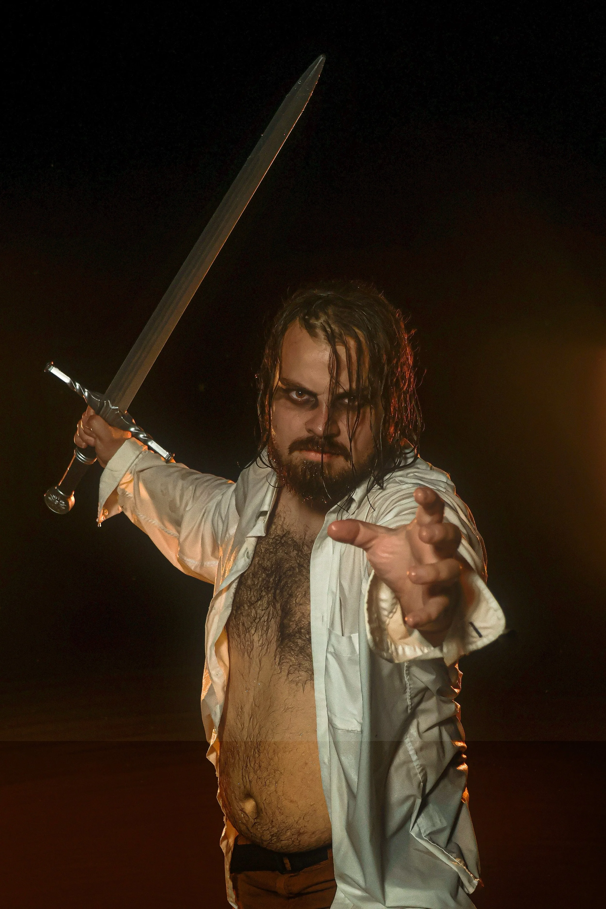 A man with long, wet hair and a beard, wearing an open white shirt, pointing towards the camera, and holding a sword in the air at night with dark background.
