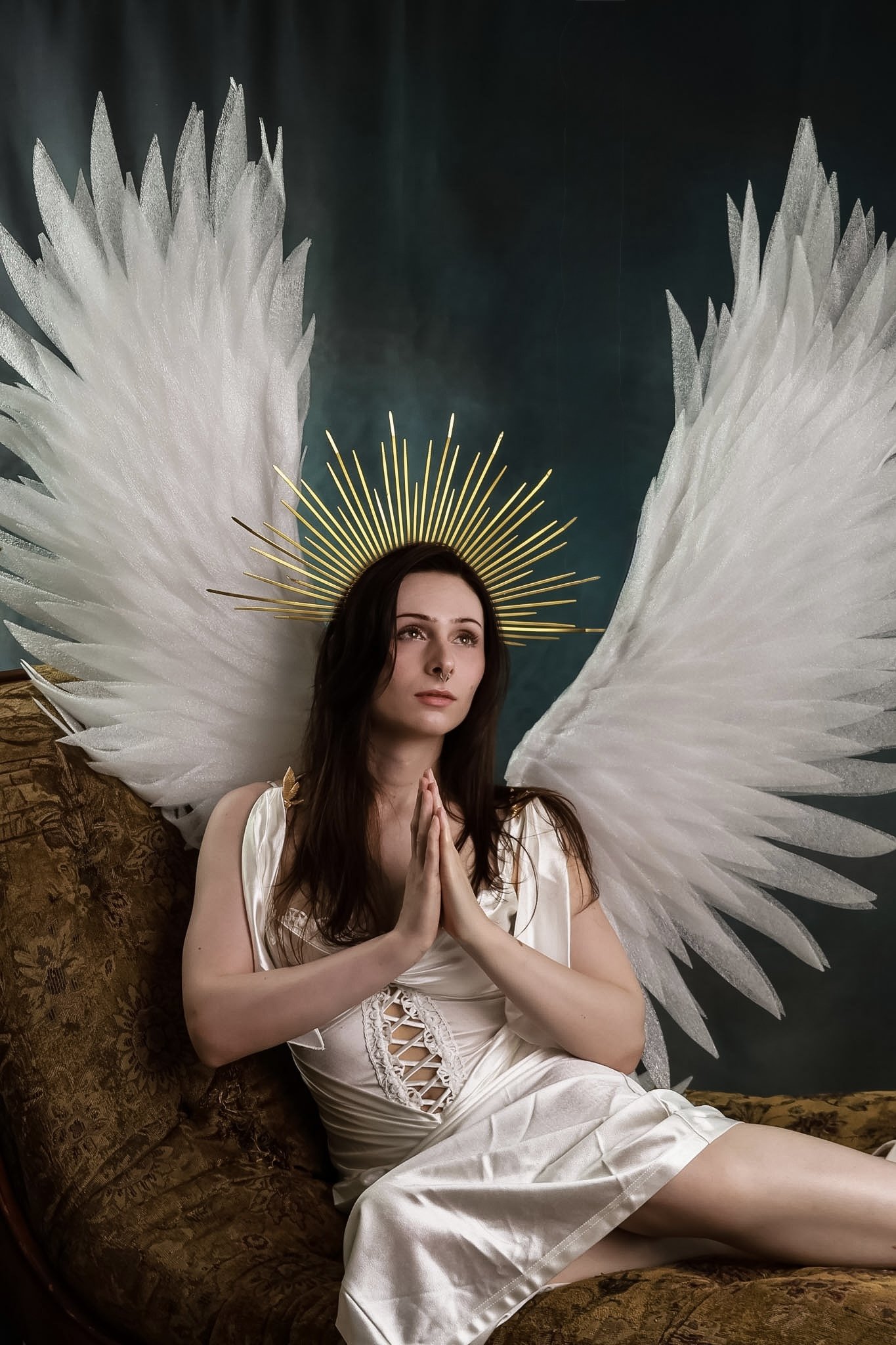 A woman with long brown hair and fair skin, dressed in a white silk-like dress, sitting on a dark ornate sofa with a floral pattern. She has her hands clasped in front of her chest in a prayer position. Behind her, there is a decorative halo-like hea