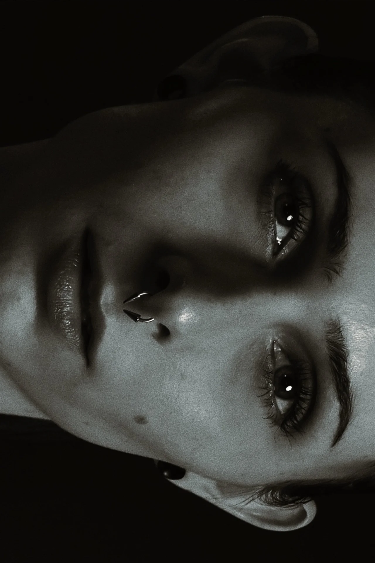 Close-up of a woman's face in black and white, with her eyes closed, her hand near her lips, and she is wearing a septum piercing.