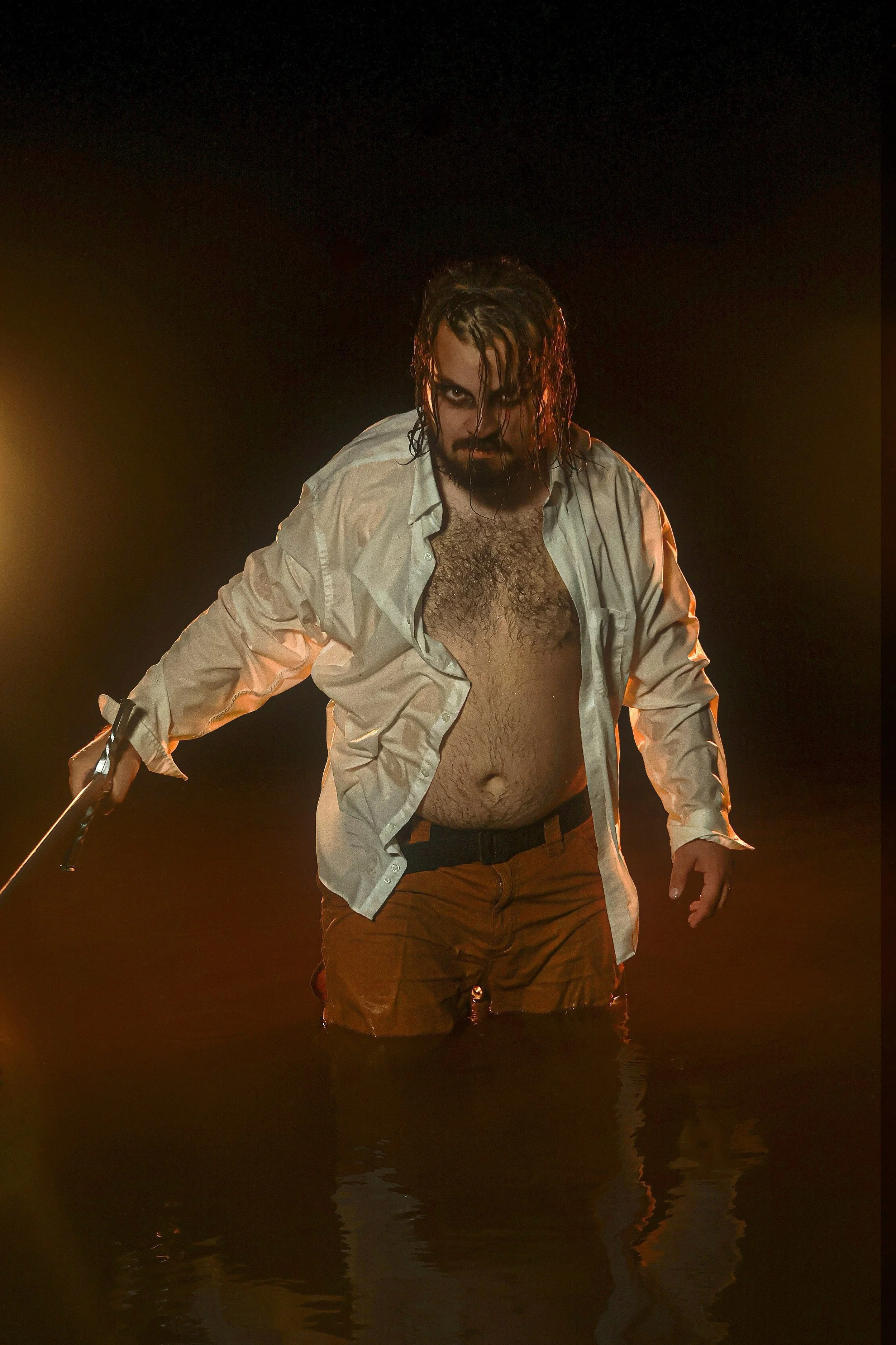 A man with long wet hair and heavy eye makeup stands in dark water, holding a gun, wearing an open white shirt and brown pants, with dramatic lighting highlighting him against a dark background.