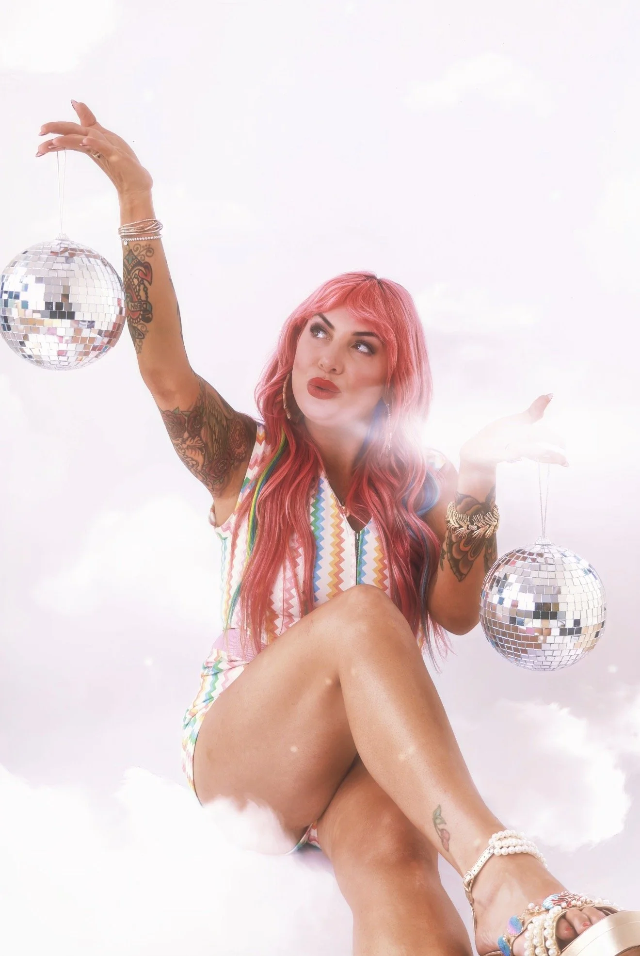 A woman with pink hair and tattoos poses with two disco balls against a white background.