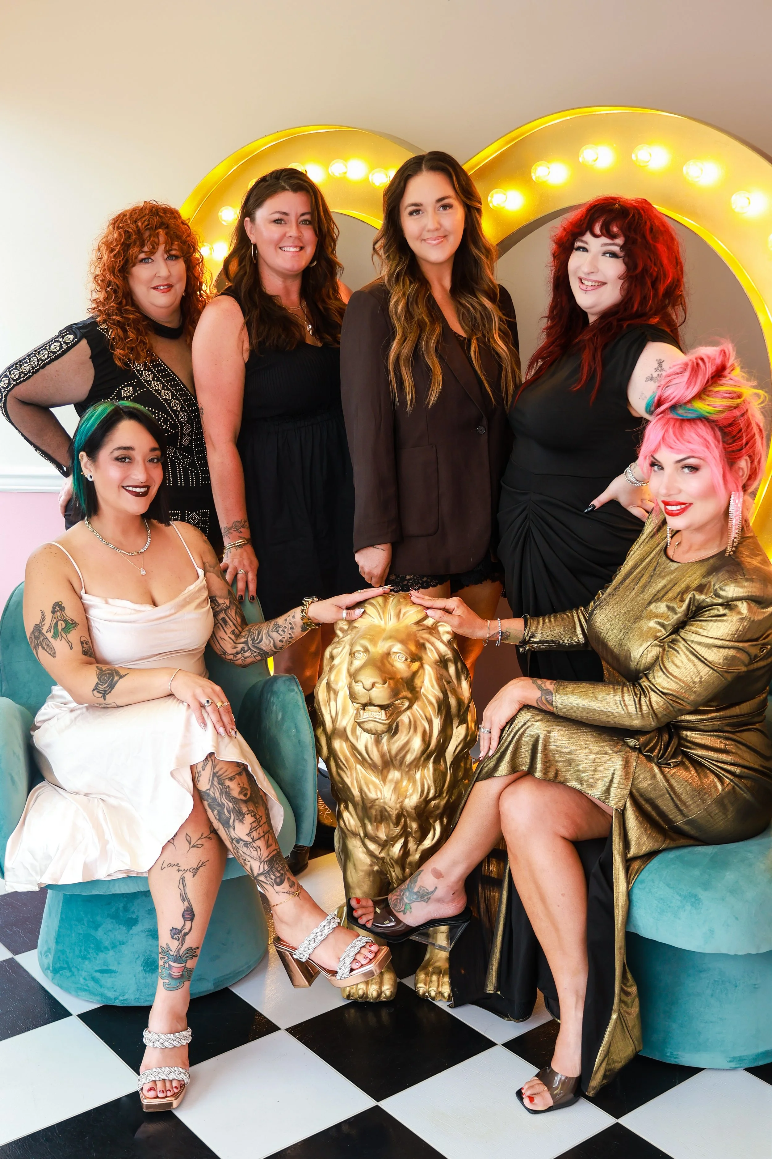 Group of six women with colorful hairstyles and tattoos, sitting and standing around a gold lion statue, posing for a photo in front of a bright yellow marquee sign with light bulbs, on a black and white checkered floor.
