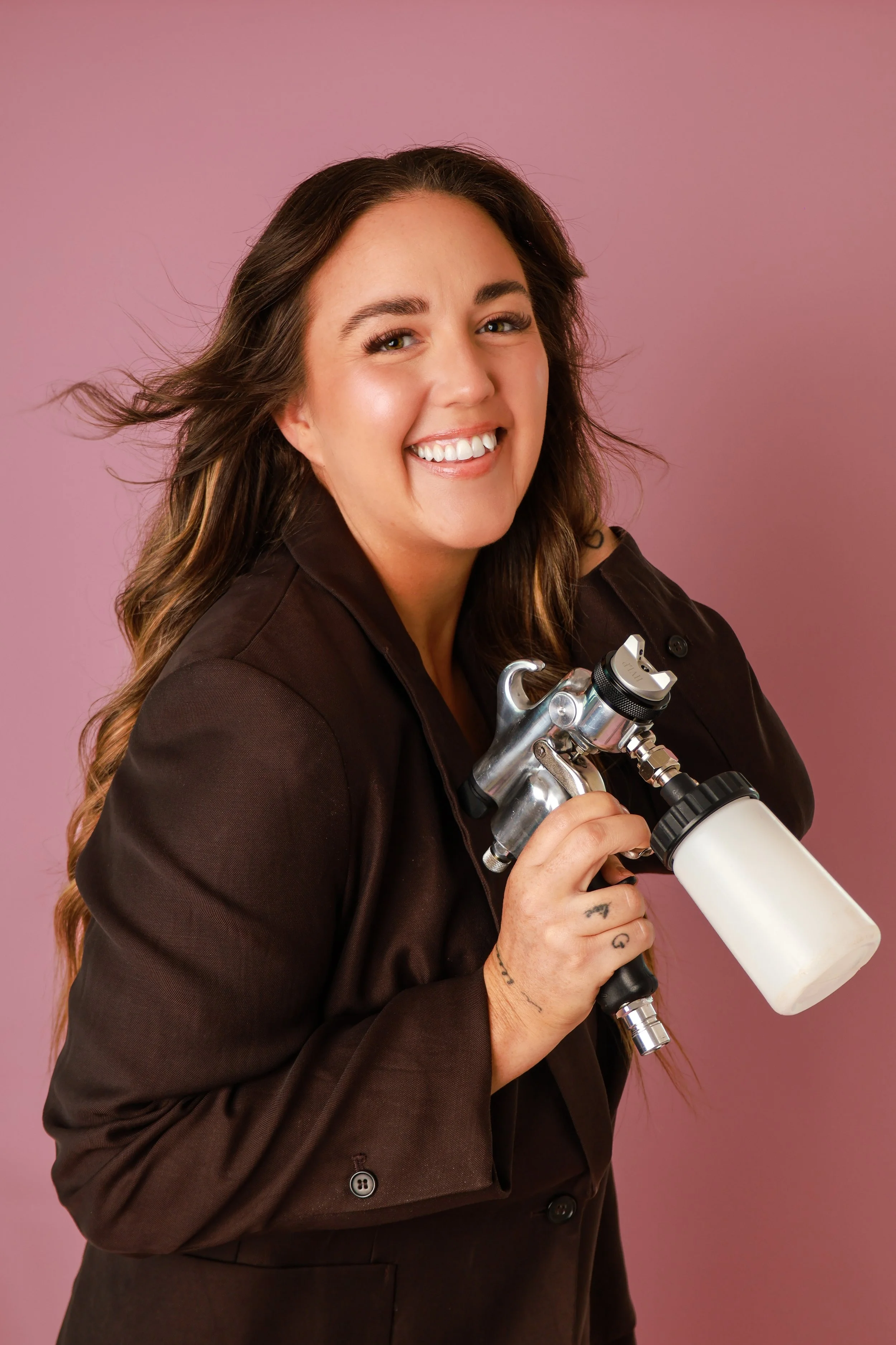 A woman with long wavy brown hair and a big smile holding a telescope, wearing a dark jacket, against a pink background.