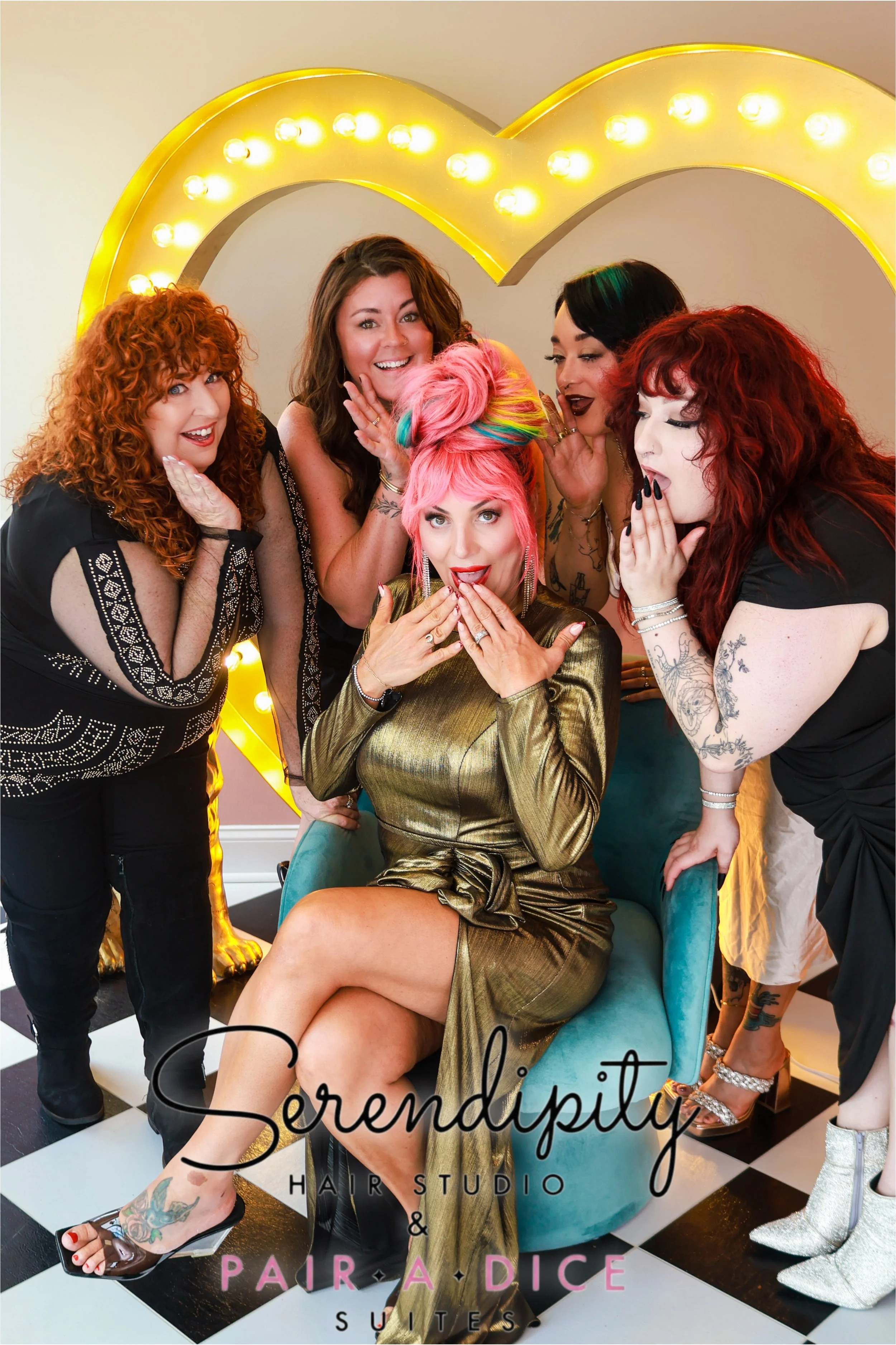 A woman with pink and rainbow-colored hair sitting on a teal chair, surrounded by four women with red, black, and curly hair, posing and whispering, with a large yellow illuminated letter and the words 'Serendipity Hair Studio & Suite' and 'Pair a Di