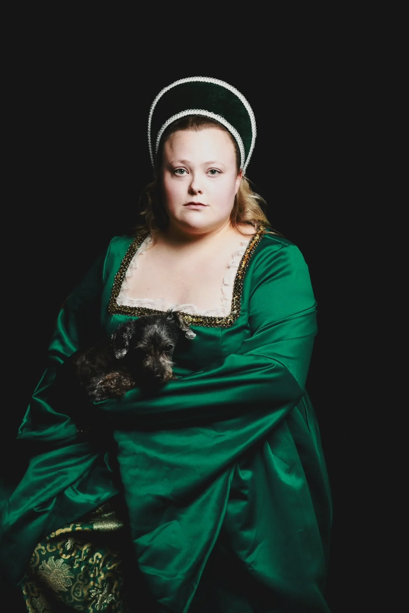 A woman dressed in an ornate green gown with gold accents and a lace neckline, wearing a tall, decorated headdress with white trim, holds a small black and gray puppy against a black background.