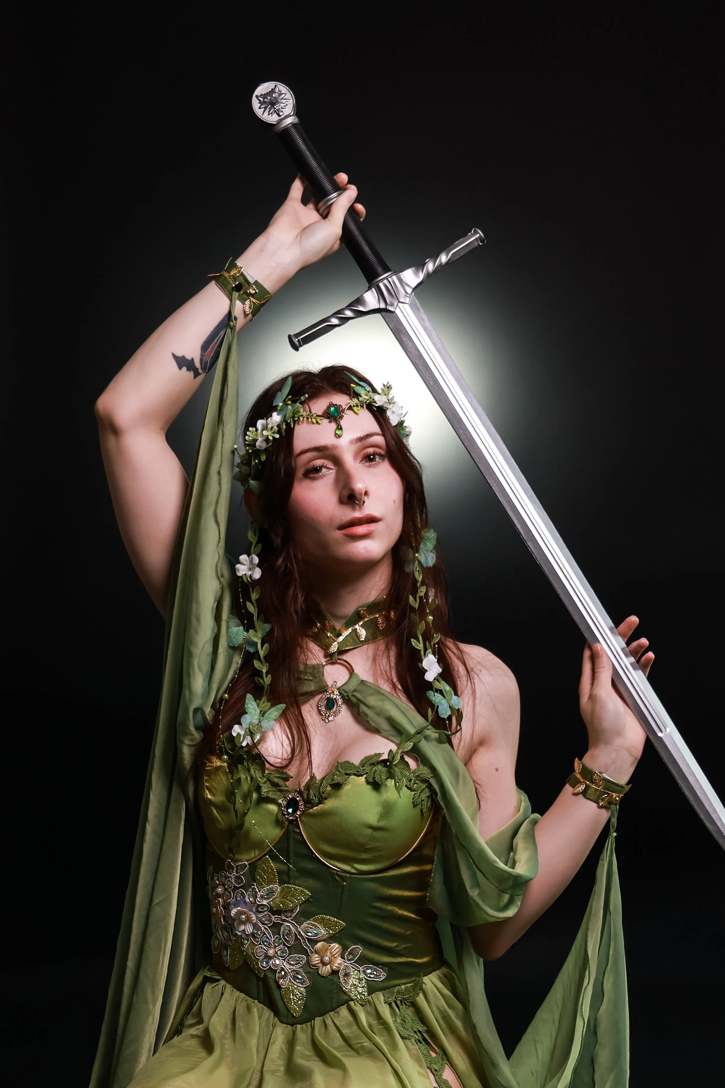 A woman with long brown hair dressed in a green fantasy costume with floral decorations and jewelry, holding a large sword with the tip pointed upwards against a dark background.