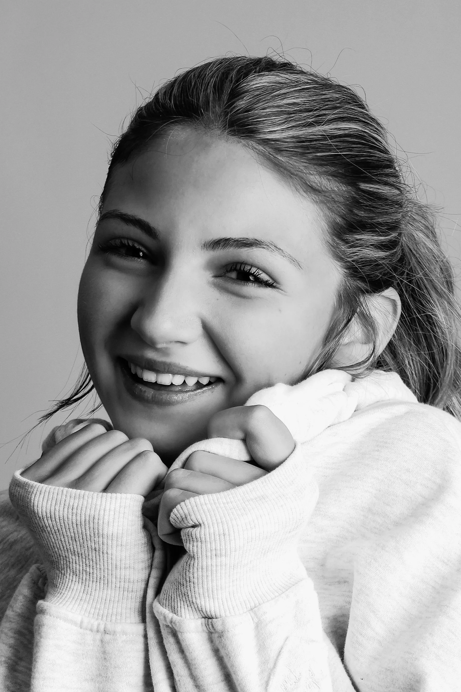 Black and white photo of a smiling young woman with her hair pulled back, wearing a hoodie, with her hands near her chin against a plain background.