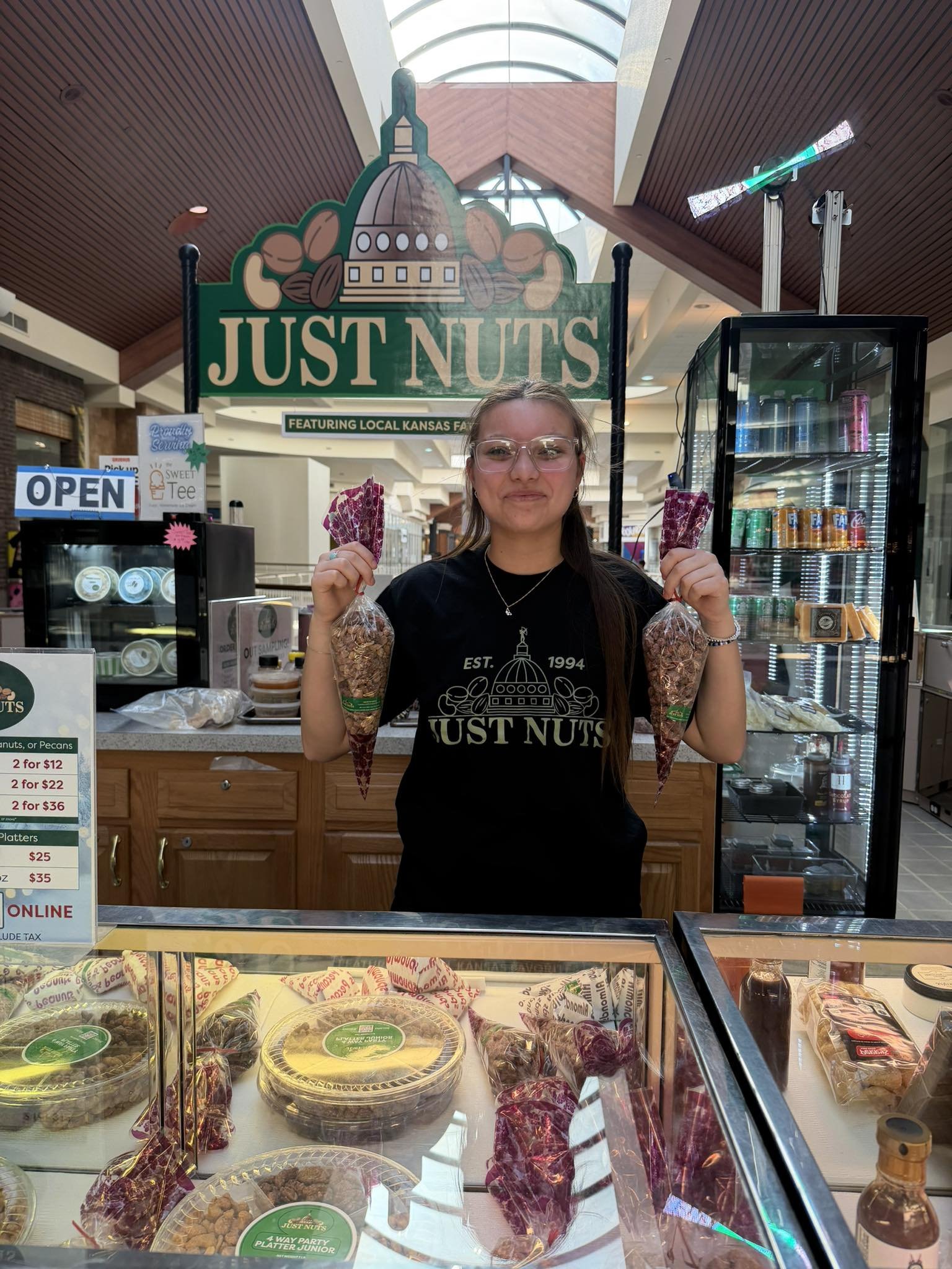 A woman with glasses standing behind a nut and snack display at a store called 'Just Nuts'. She is holding small bags of nuts, with various packaged nuts and snacks visible in front of her.