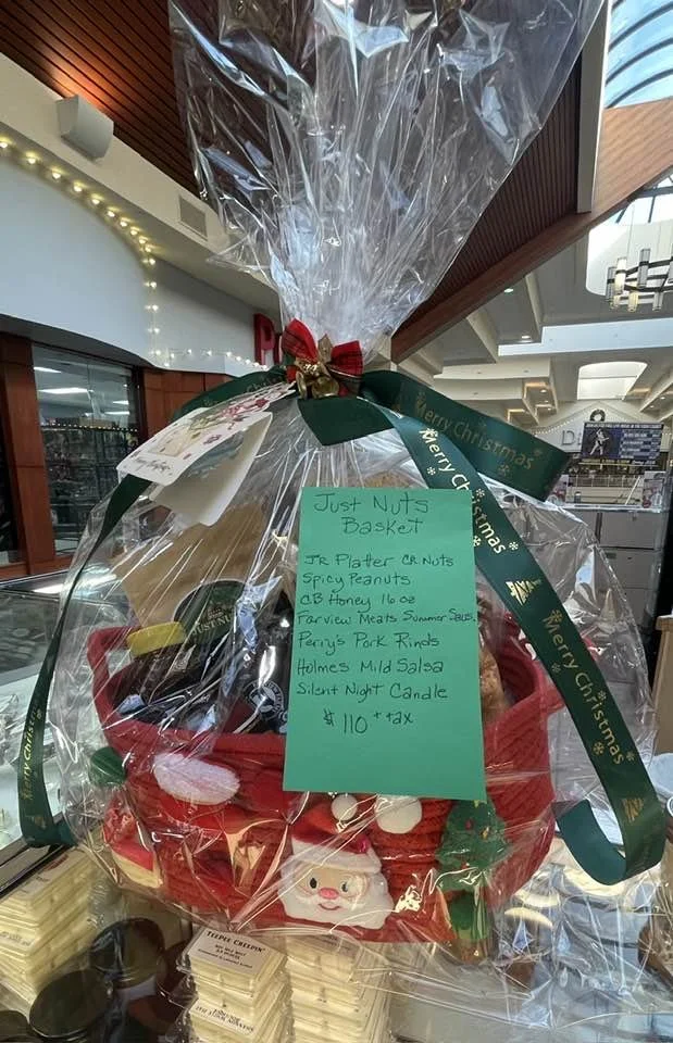 Christmas gift basket wrapped in clear plastic with a green ribbon and red bow, containing snack items, a Santa Claus plush, and a handwritten note listing contents and price.