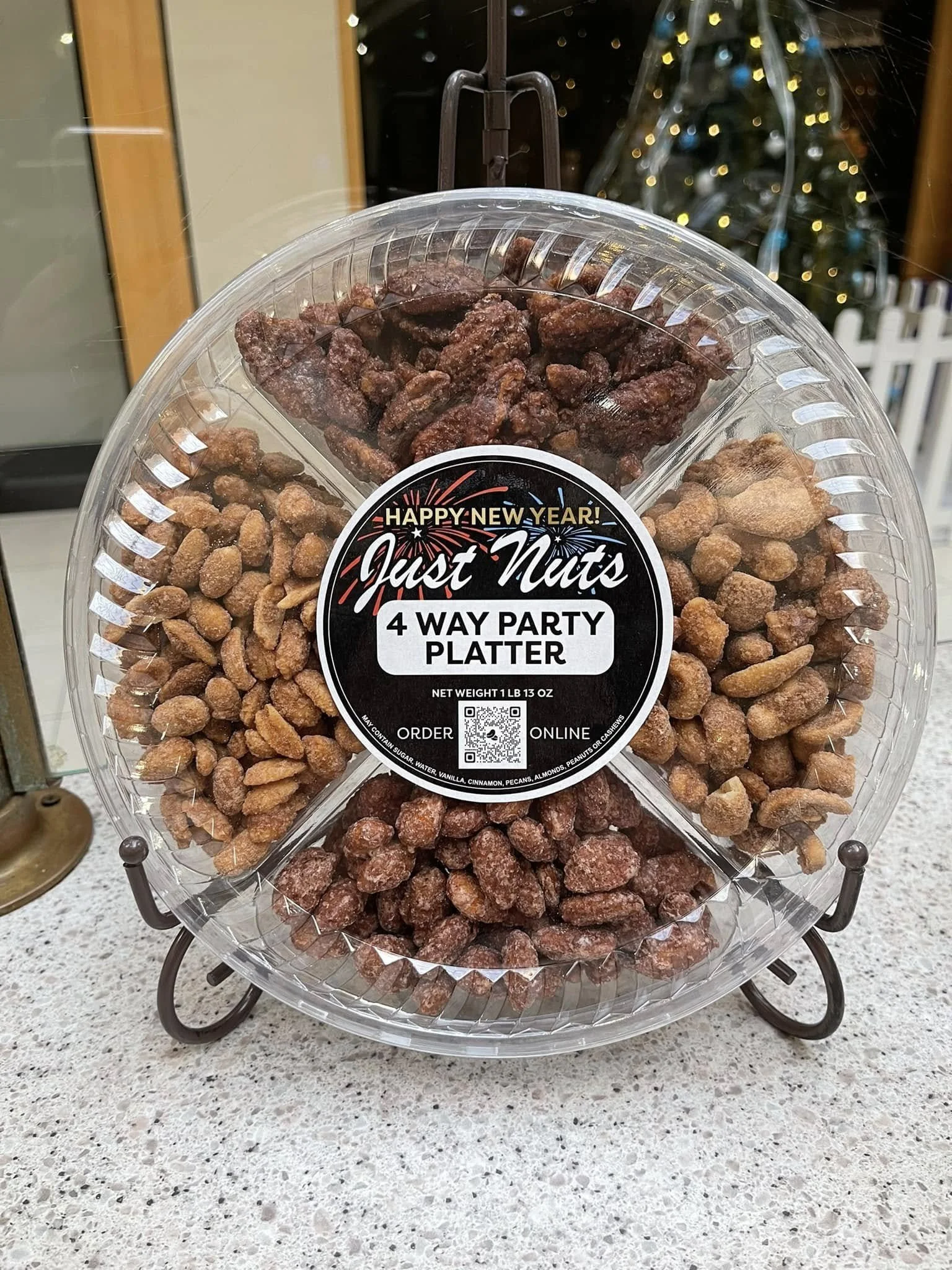 Clear plastic tray with assorted flavored nuts, including caramelized, chocolate, and spicy nuts, with a "Happy New Year!" and "Just Nuts" label, placed on a small stand on a kitchen countertop, with a Christmas tree in the background.