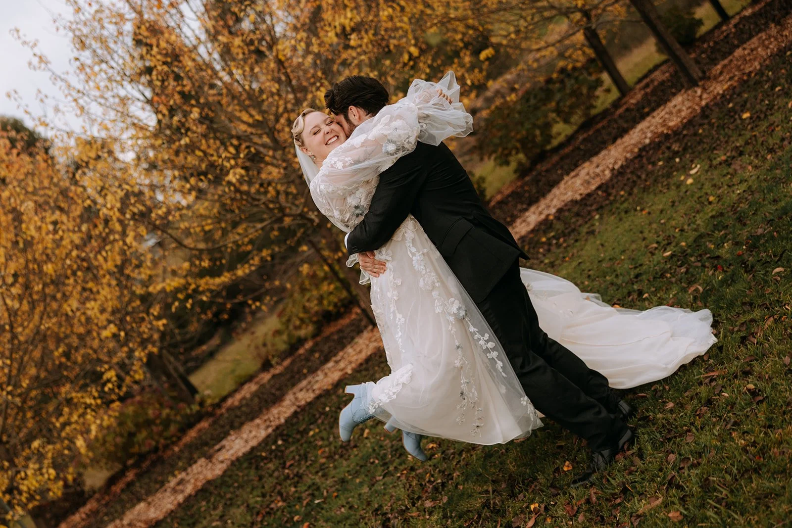 A smiling bride and groom happily dancing outdoors during fall, surrounded by orange and yellow trees, with fallen leaves on the ground.