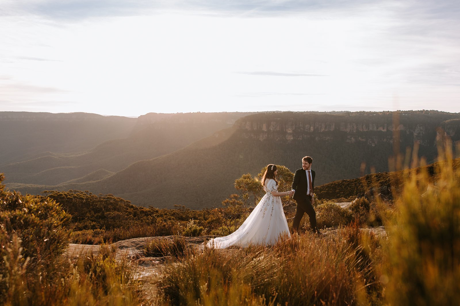 A bride and groom holding hands and walking through a scenic mountainous landscape during sunset.