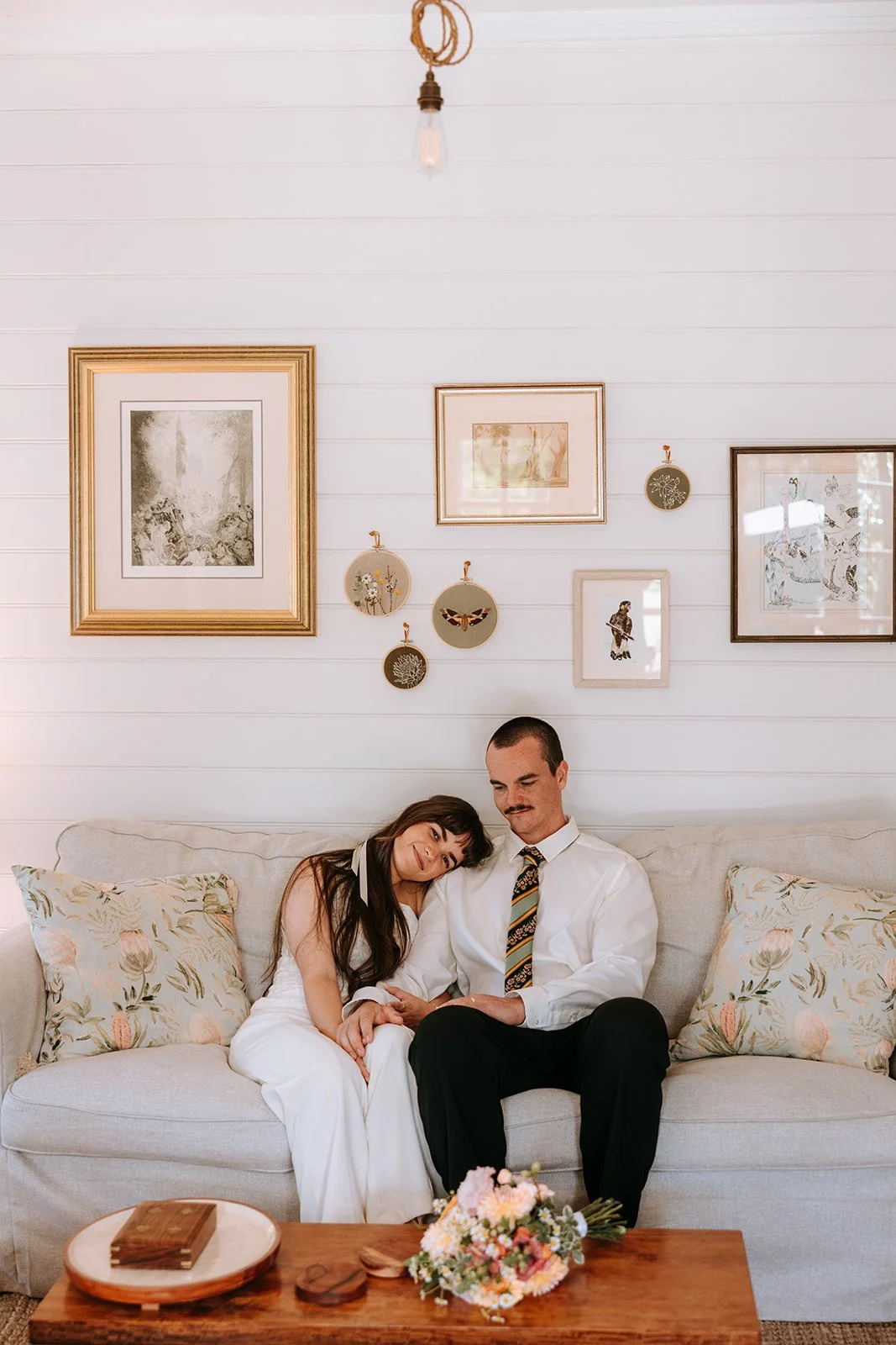 A couple sitting on a beige couch, with the woman resting her head on the man's shoulder, holding hands. The woman wears a white dress, and the man wears a white shirt and patterned tie. The wall behind them features framed artwork and hanging decorative plates, with a wooden coffee table with flowers and books in front.