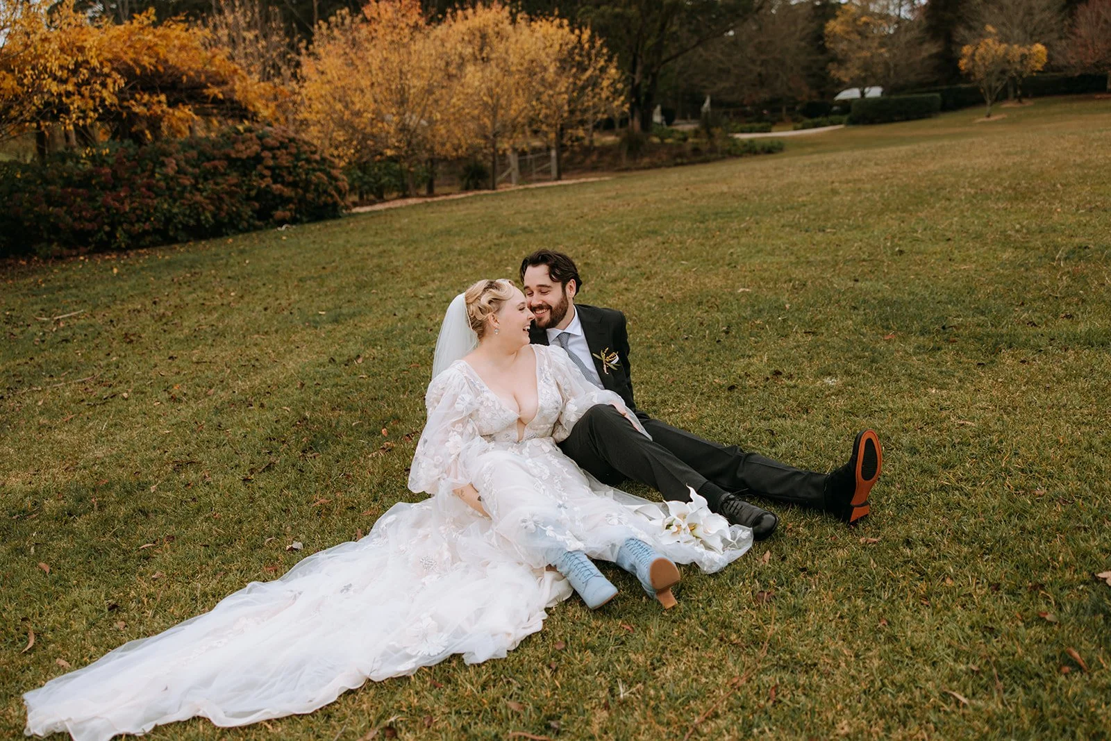 A bride and groom sitting on grass in a park during autumn, smiling and laughing together. The bride wears a white wedding dress with floral embroidery and a veil, and the groom wears a dark suit with a tie. The background has colorful fall foliage.