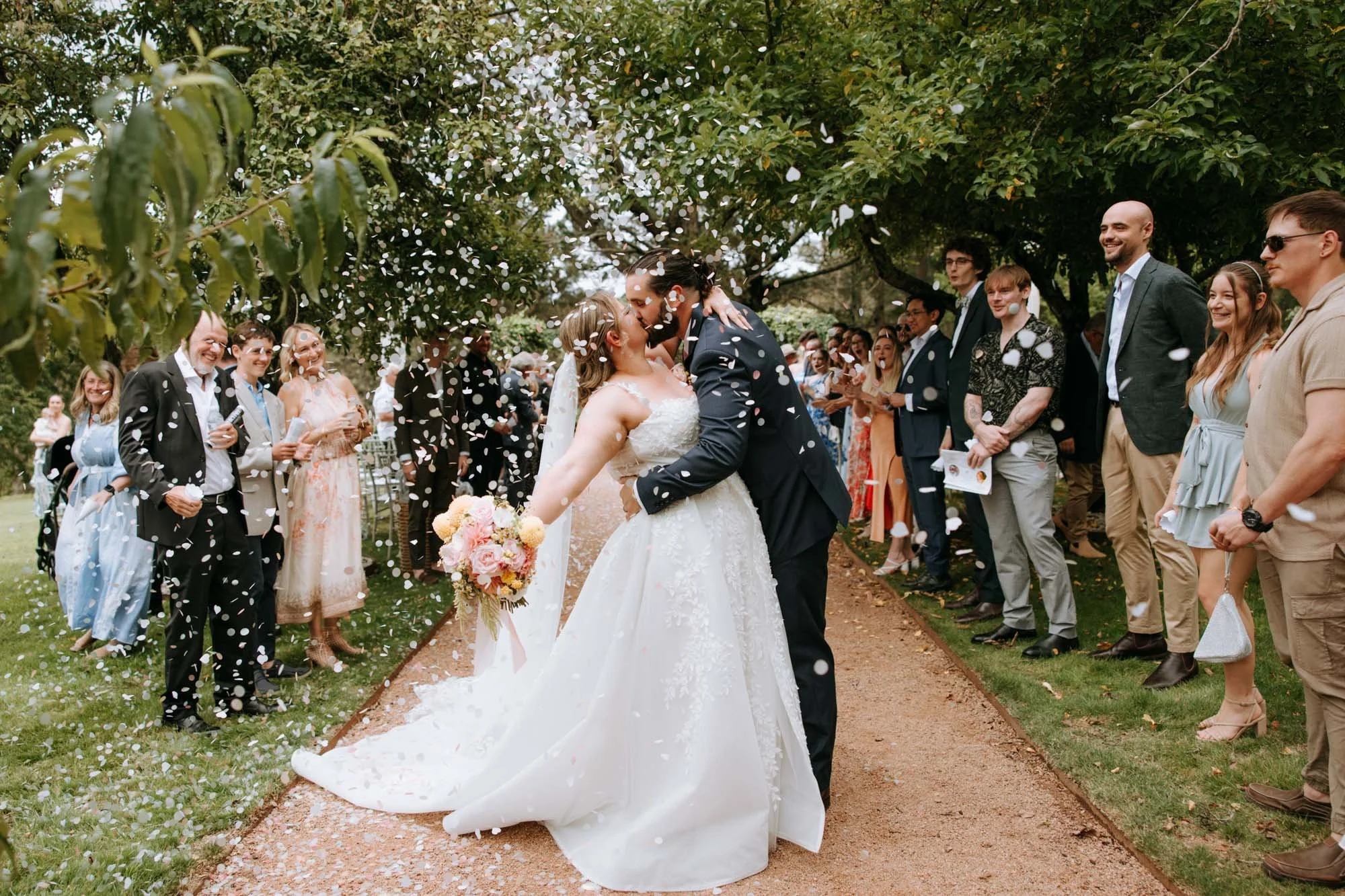 A bride and groom share a kiss under a tree at their outdoor wedding, surrounded by friends and family celebrating with white flower petals falling.