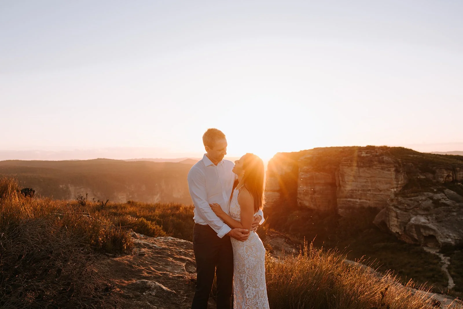 A couple stands close together on a rocky hillside during sunset, with a canyon and large rock formations in the background, illuminated by the setting sun.