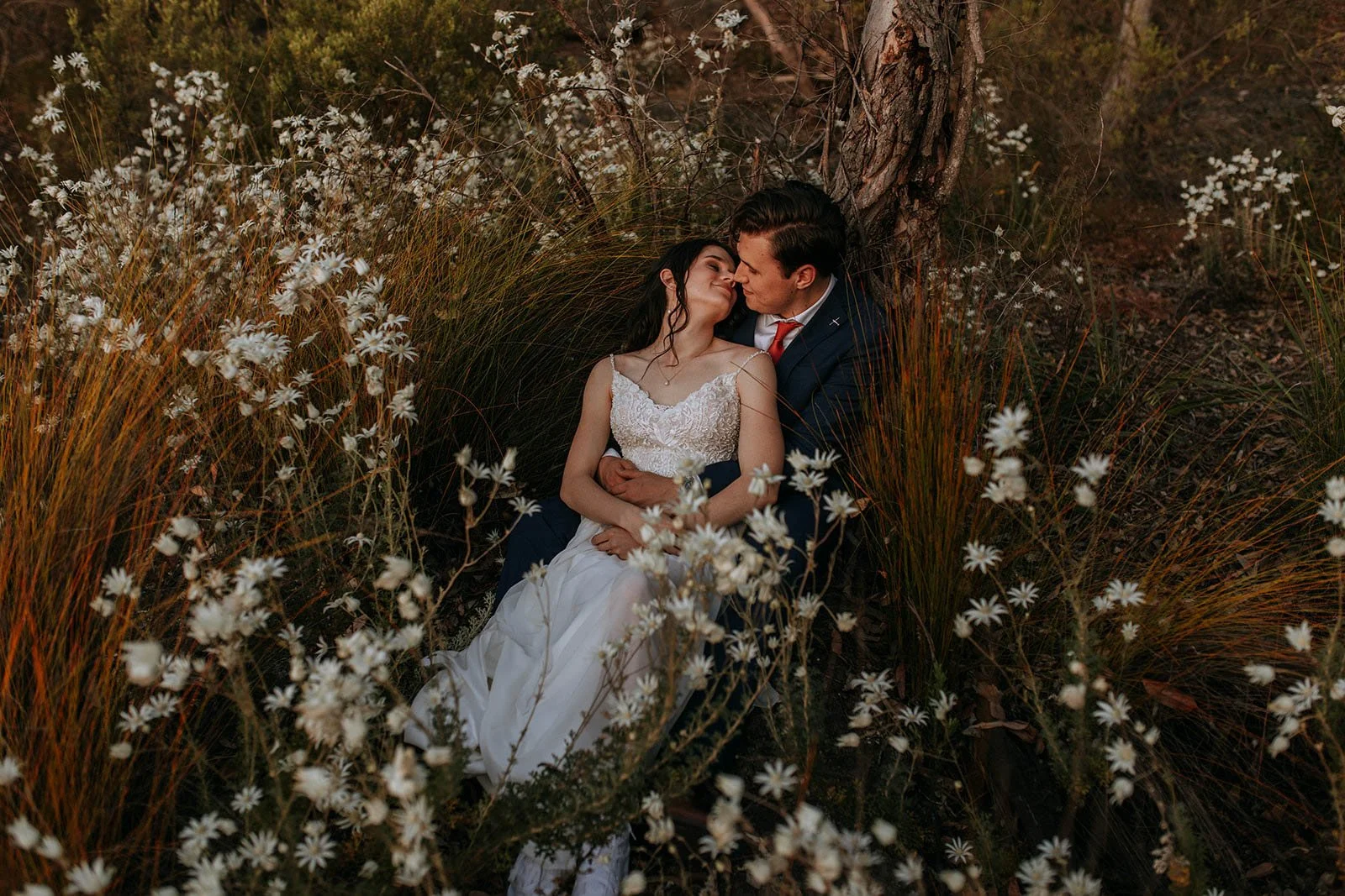 A couple dressed in wedding attire sitting close together in a natural outdoor setting surrounded by white wildflowers and tall grasses, with a tree in the background, sharing an intimate moment.
