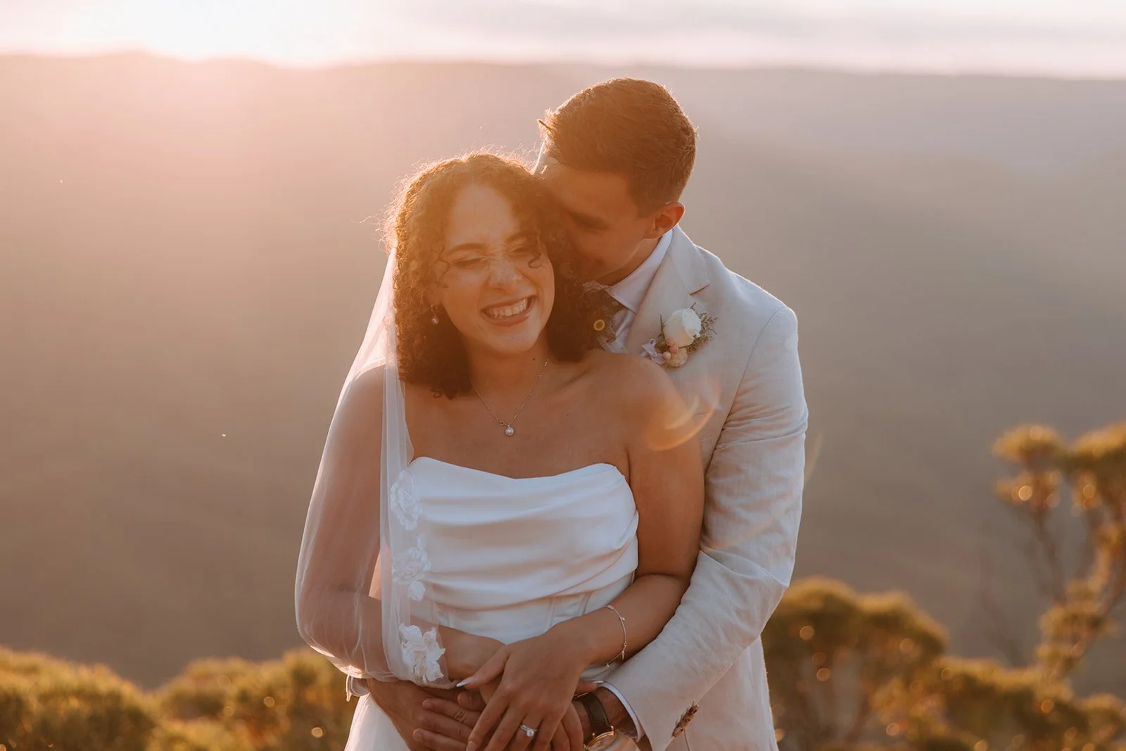A bride and groom embrace outdoors during sunset, with the groom kissing the bride's temple as she smiles. The bride is wearing a strapless white wedding dress and the groom is in a white suit with a boutonniere. There are trees and mountains in the background, bathed in warm sunlight.