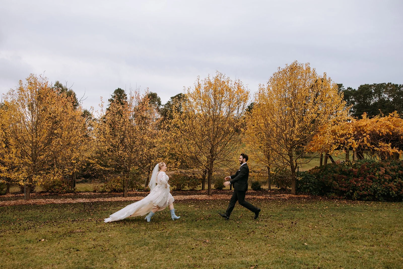 A bride and groom are in a park with fall foliage, running towards each other and smiling, with trees and bushes in the background.