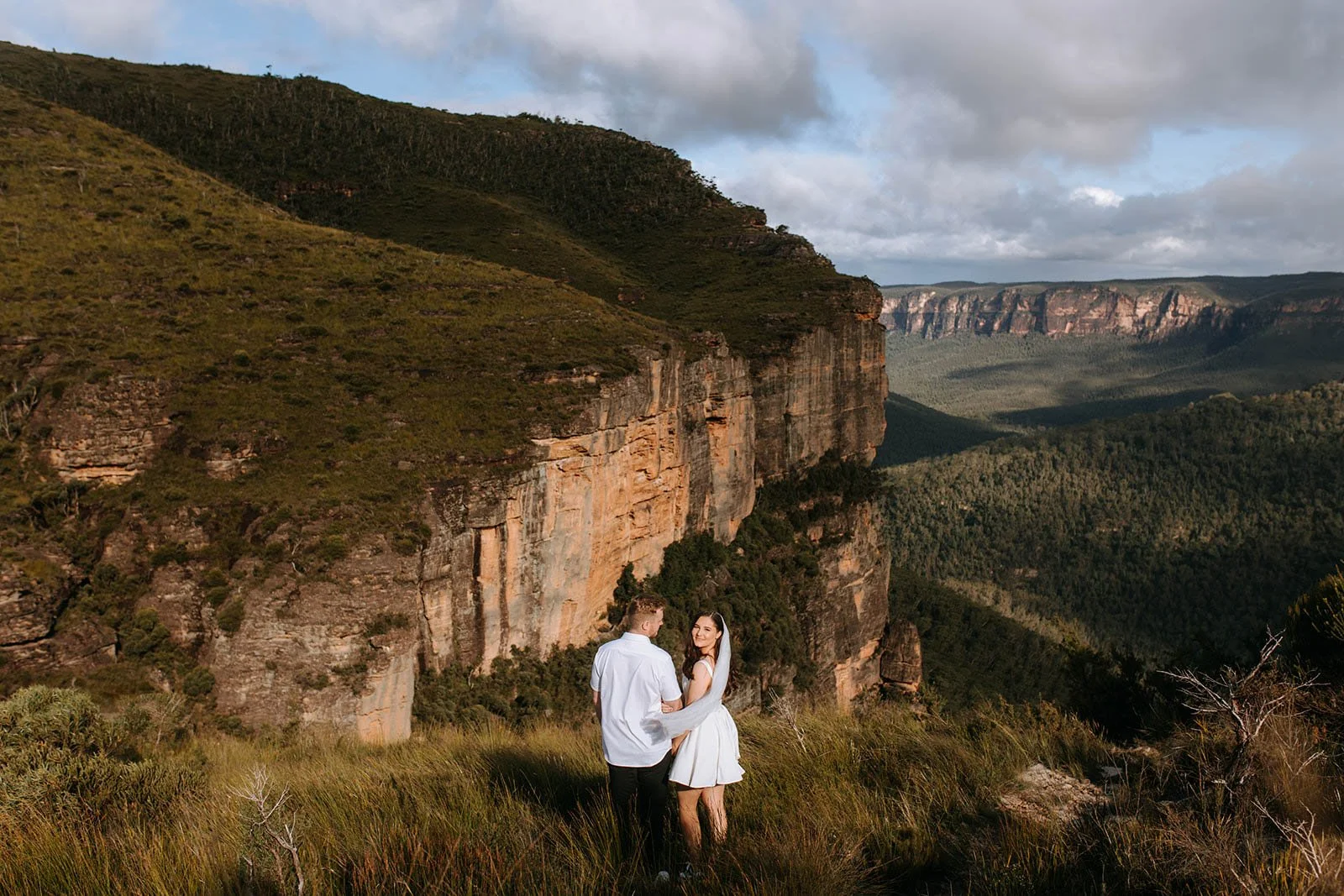 A couple dressed in wedding attire standing on a grassy hilltop with a rock formation and a canyon in the background under a cloudy sky.