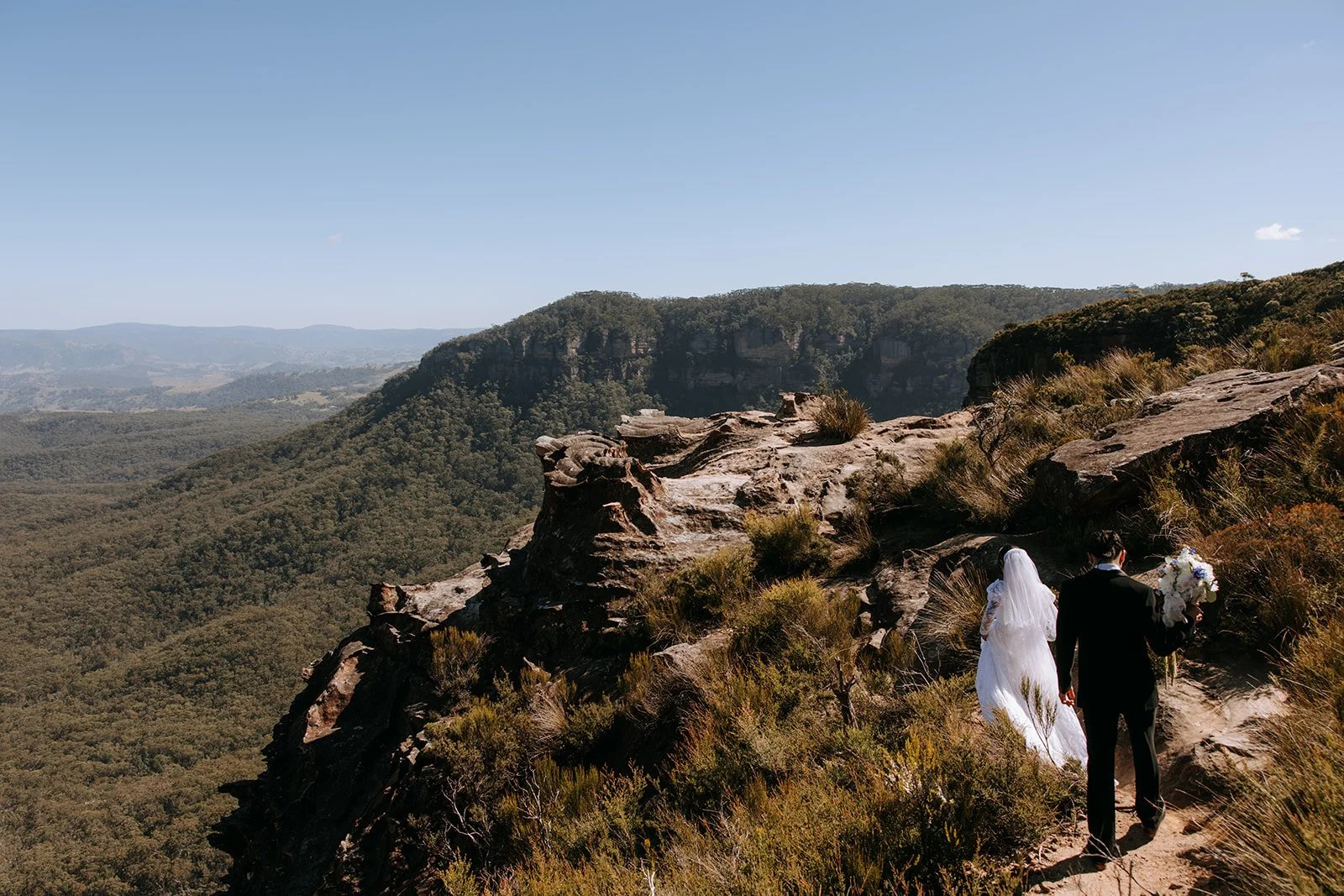 A bride and groom walk hand in hand along a rocky mountain trail overlooking a vast valley and forest under a clear blue sky, with the groom holding a bouquet of white flowers.