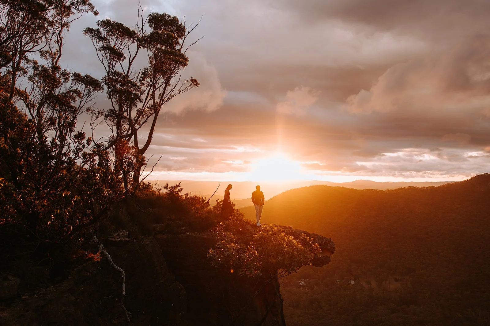 People standing on a cliff at sunset, surrounded by trees and rolling hills, with dramatic clouds in the sky.