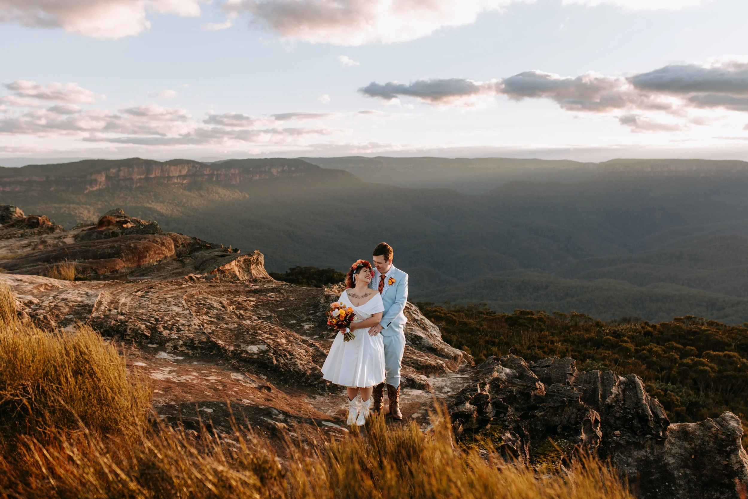 A bride and groom standing on a rocky landscape with a vast canyon and cloudy sky in the background, during sunset.