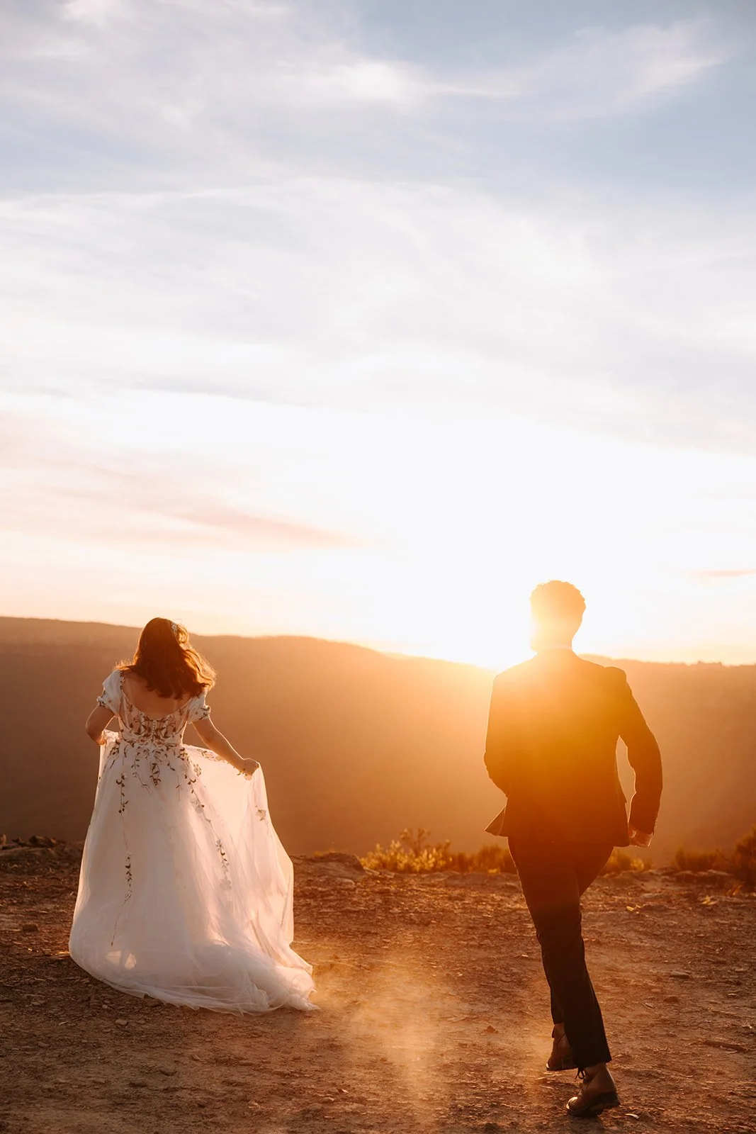 A couple in wedding attire, a woman in a white dress and a man in a suit, walking on a dirt path during sunset with mountains in the background.