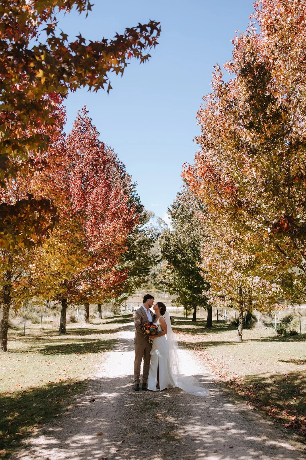 A bride and groom standing close together on a dirt path surrounded by colorful fall trees, holding a bouquet and sharing a kiss under a clear blue sky.