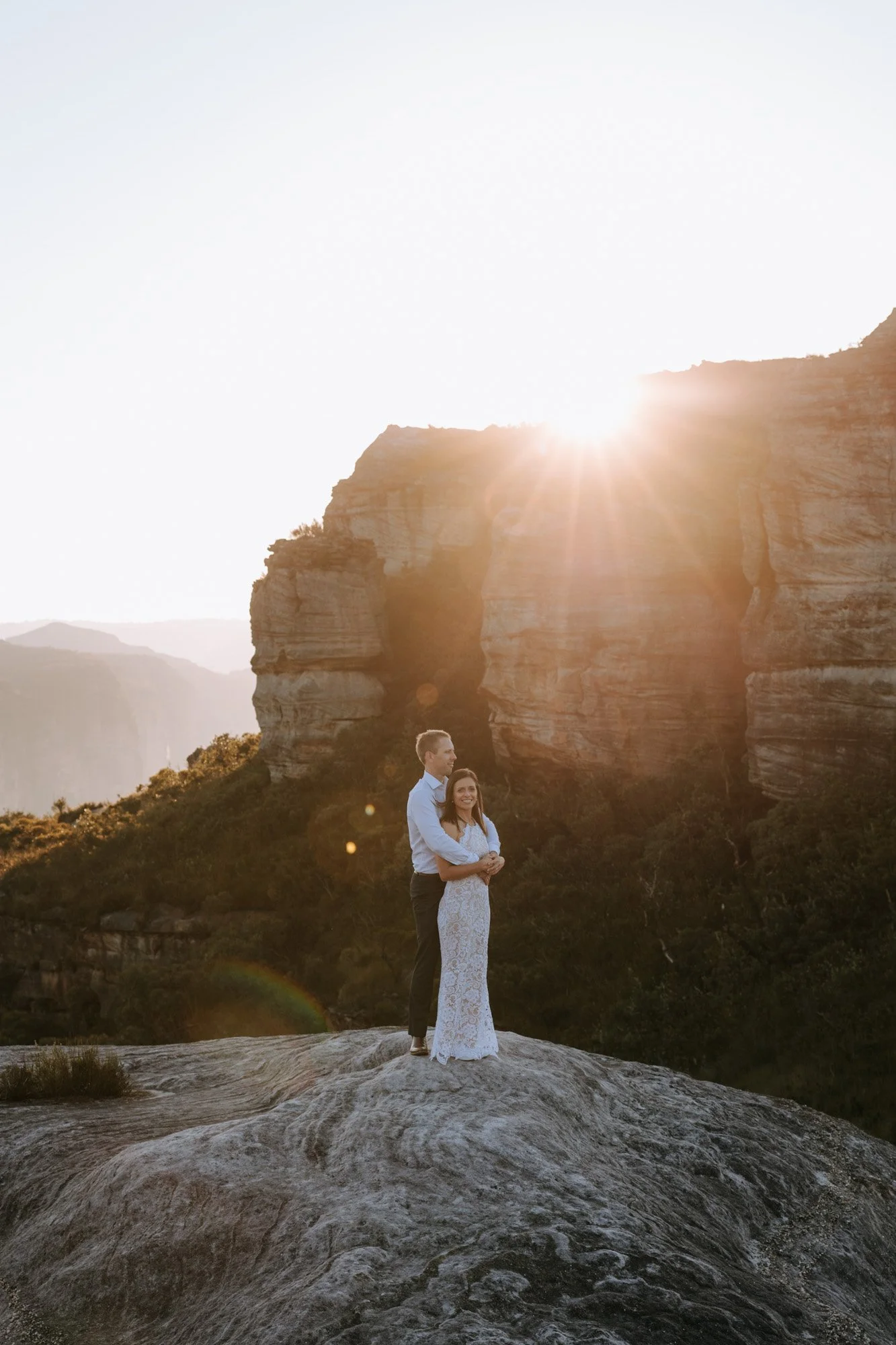 A couple standing on a large rock in a scenic outdoor landscape during sunset, with a rocky formation in the background and the sun setting behind the rocks.