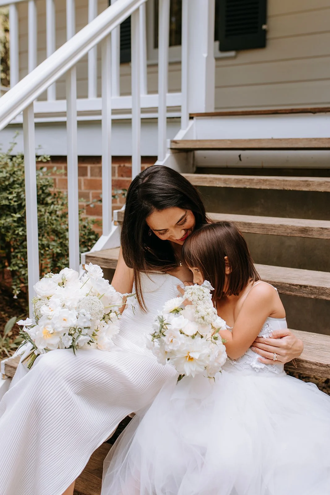 A woman in a white dress and a young girl in a white dress sitting on wooden stairs outside, hugging and looking down at white flower bouquets.