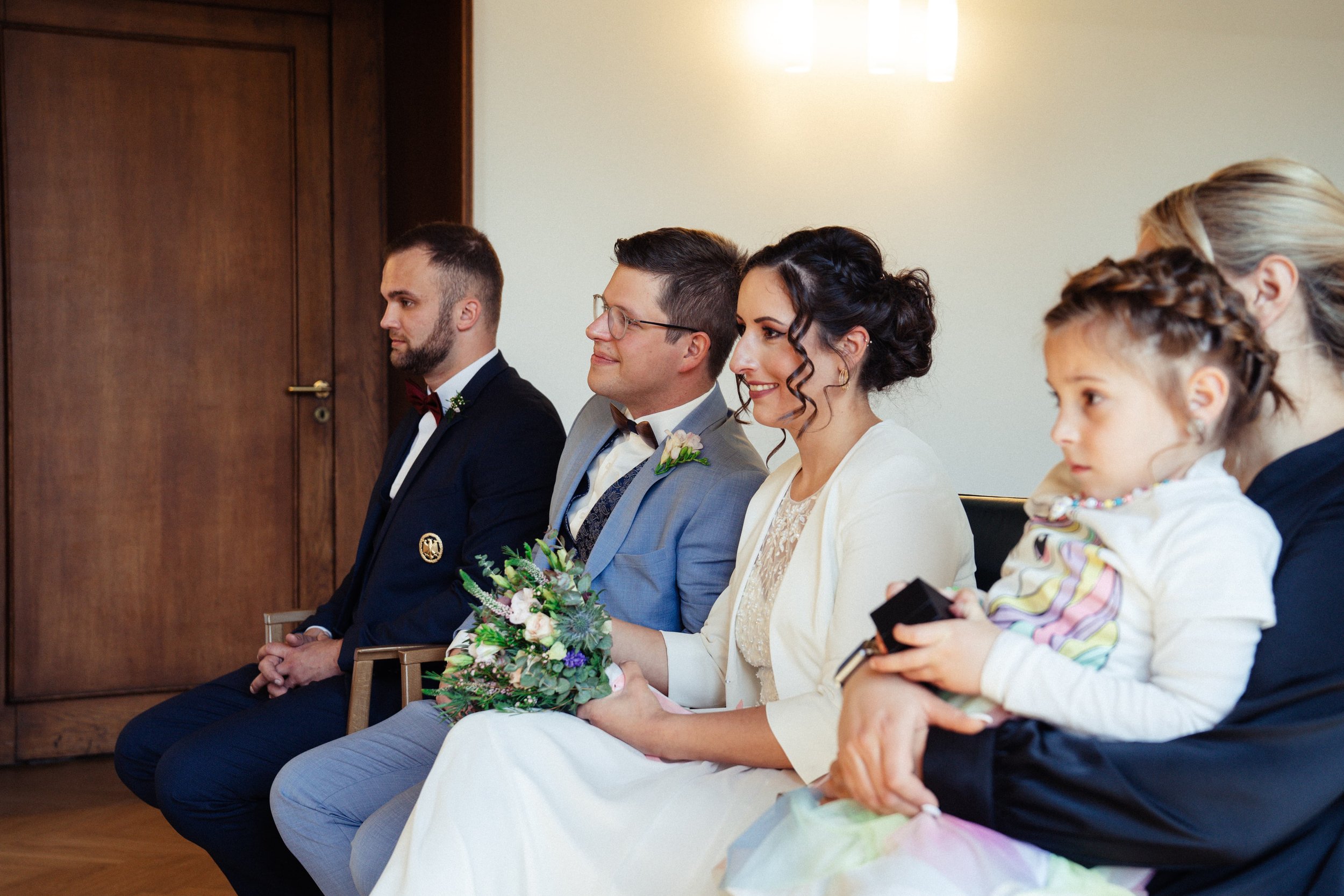 Eine Gruppe von Menschen sitzt in einer Reihe bei einer Hochzeitszeremonie, darunter ein Bräutigam, eine Braut, ein Mann und Kinder, vermutlich während einer Trauung im Innenraum.