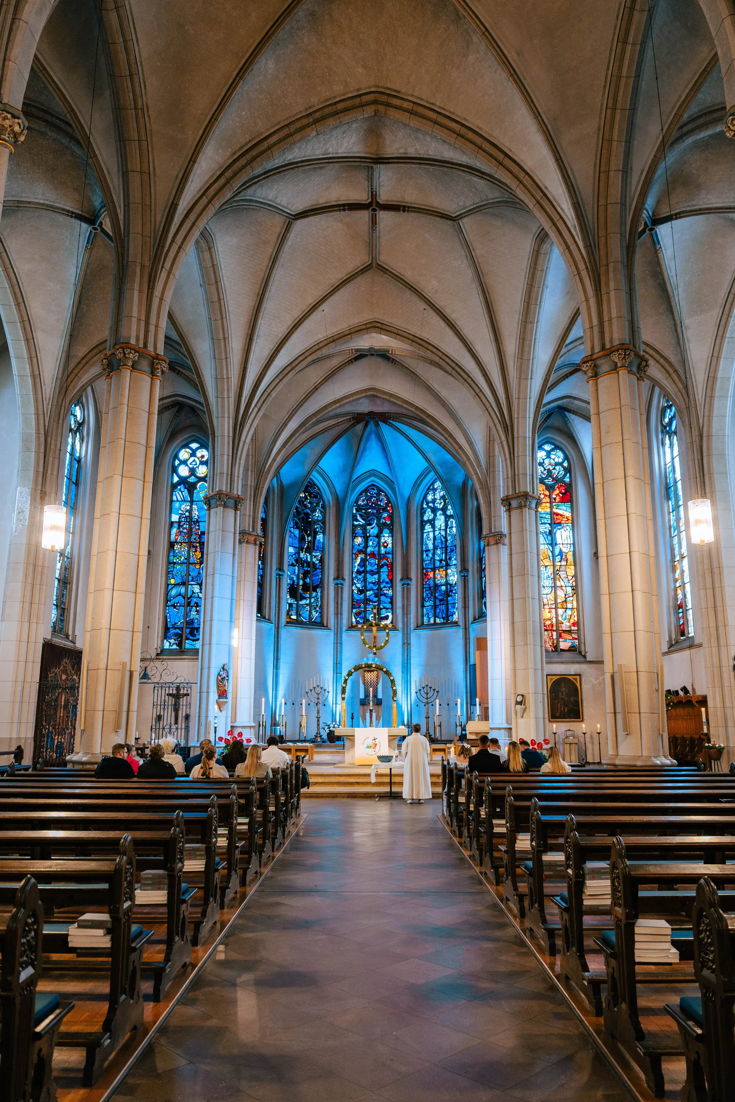 Innenansicht einer gotischen Kirche mit hohen, gewölbten Decken und bunten Glasfenstern. Im Vordergrund sind Kirchenbänke mit anwesenden Menschen. Im Altarbereich stehen Kerzen und religiöse Symbole, ein Priester steht vor den Gläubigen.