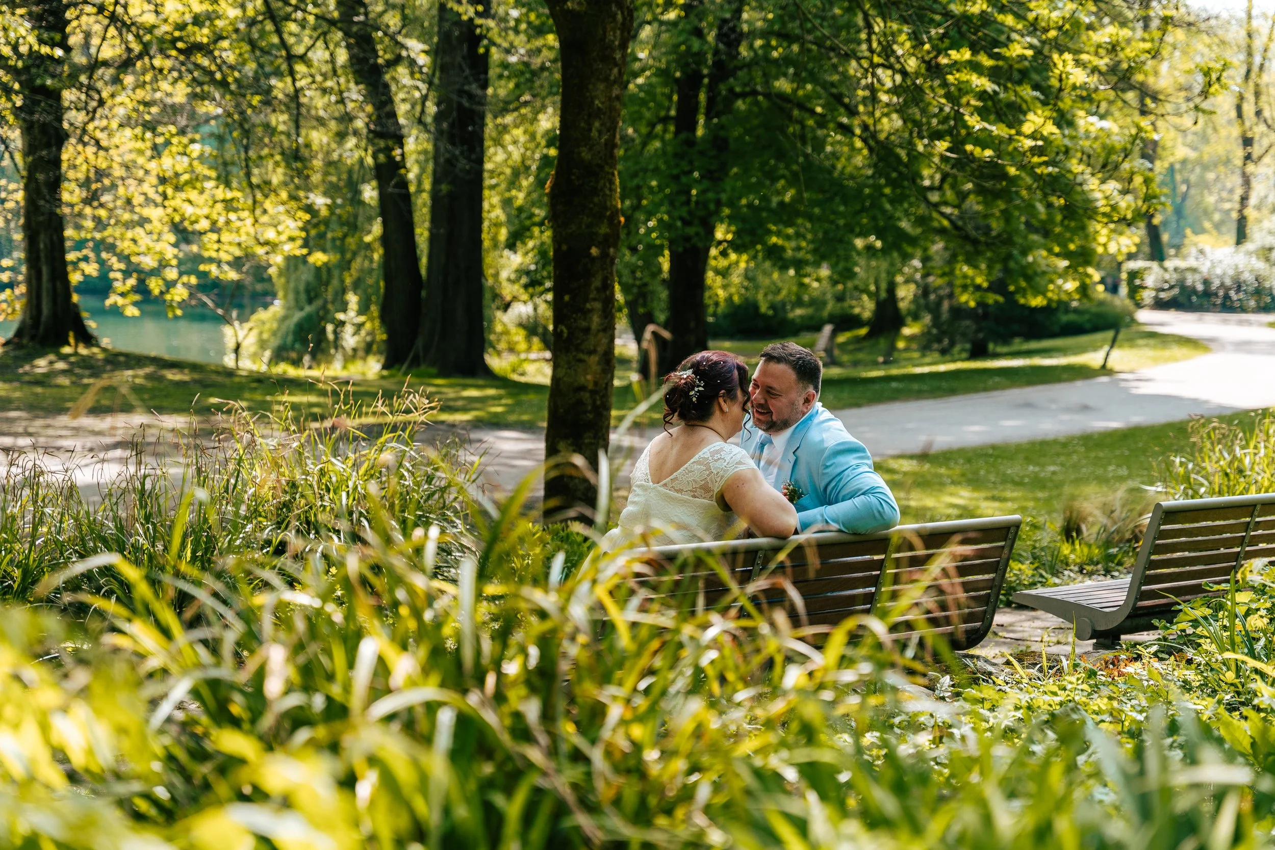 Ein lächelndes Paar sitzt auf einer Parkbank in einem grünen Park bei Sonnenlicht, umgeben von Bäumen und Pflanzen.