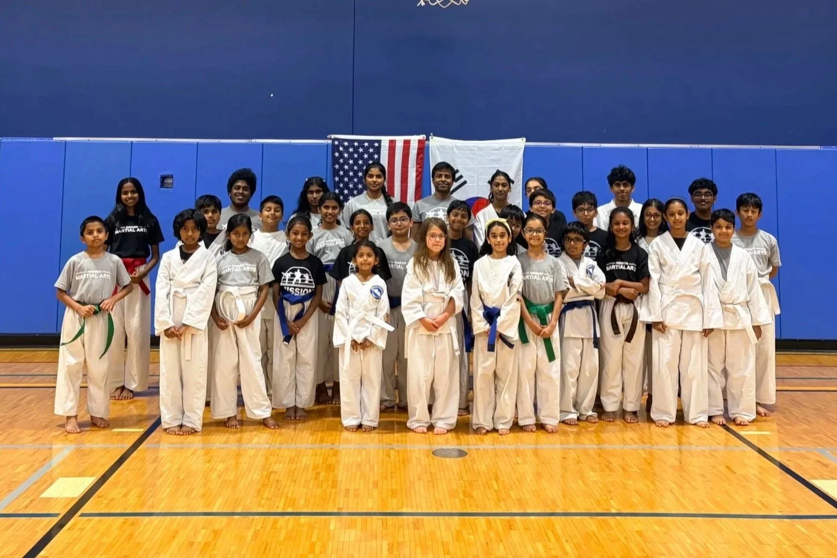 Group of children and adults in martial arts uniforms and t-shirts, posing for a group photo in a gymnasium with Blue wall and American and South Korean flags in the background.