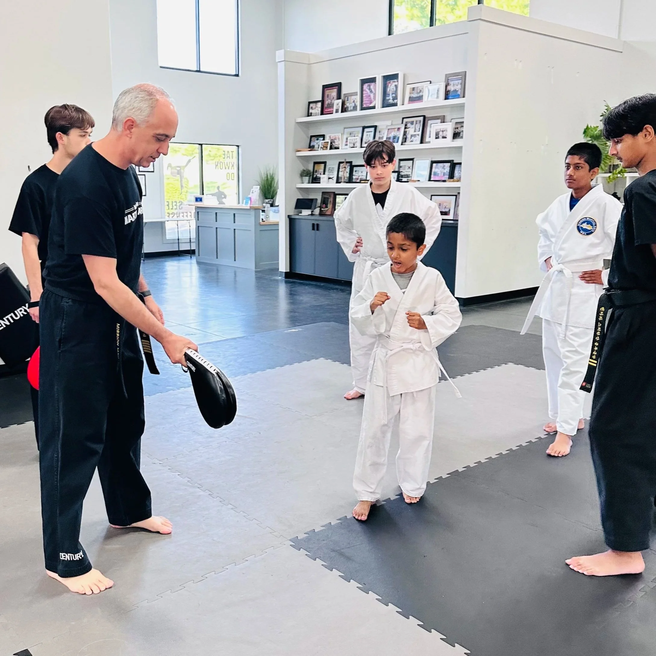 A martial arts class with children and an instructor in white uniforms practicing on a black martial arts mat in a bright room with large windows and framed photos on the wall.