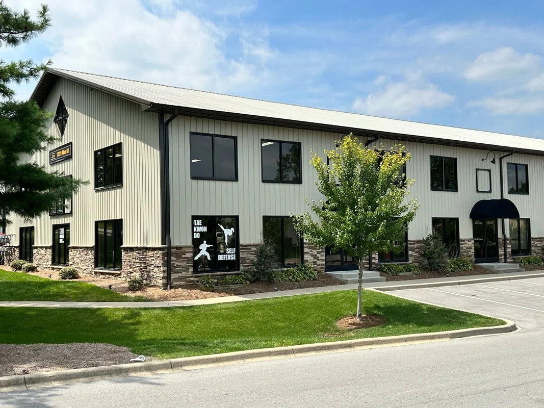 A two-story commercial building with beige siding, stone accents, and black-framed windows, featuring a Taekwondo school sign in the window stating "TAE KWON DO SELF DEFENSE," and a parking lot with green grass and a small tree in front.