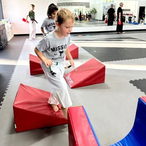 Children practicing martial arts on soft training mats in a dojo, with one child in focus stepping over red padded blocks.