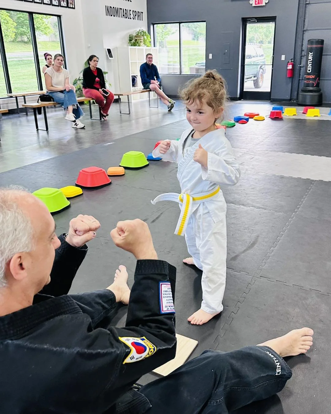 A young girl in a karate uniform with a yellow belt stands in a martial arts studio, making a fighting pose and showing a confident smile. An older man in a black gi sits on the floor in front of her with fists raised in a playful fighting stance. In the background, four people sit on benches inside the studio, watching the interaction. The studio has large windows, black mats on the floor, colorful training cones and pads, and motivational signage on the walls.