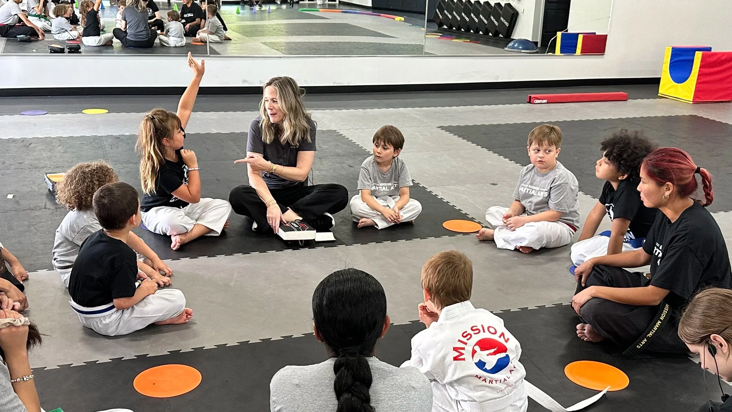 An instructor is leading a martial arts class for children seated in a circle on a mat. The instructor is gesturing with her right hand, and some children are listening attentively. The background features a wall mirror, colorful equipment, and other children practicing in the mirror.