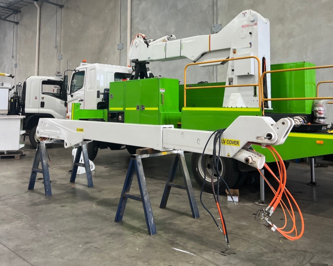 A large service truck equipped with a hydraulic arm, positioned inside a warehouse or garage. The truck has a white and green color scheme, with orange and black hydraulic hoses hanging from the arm. The truck is supported on stands for maintenance or repair work.