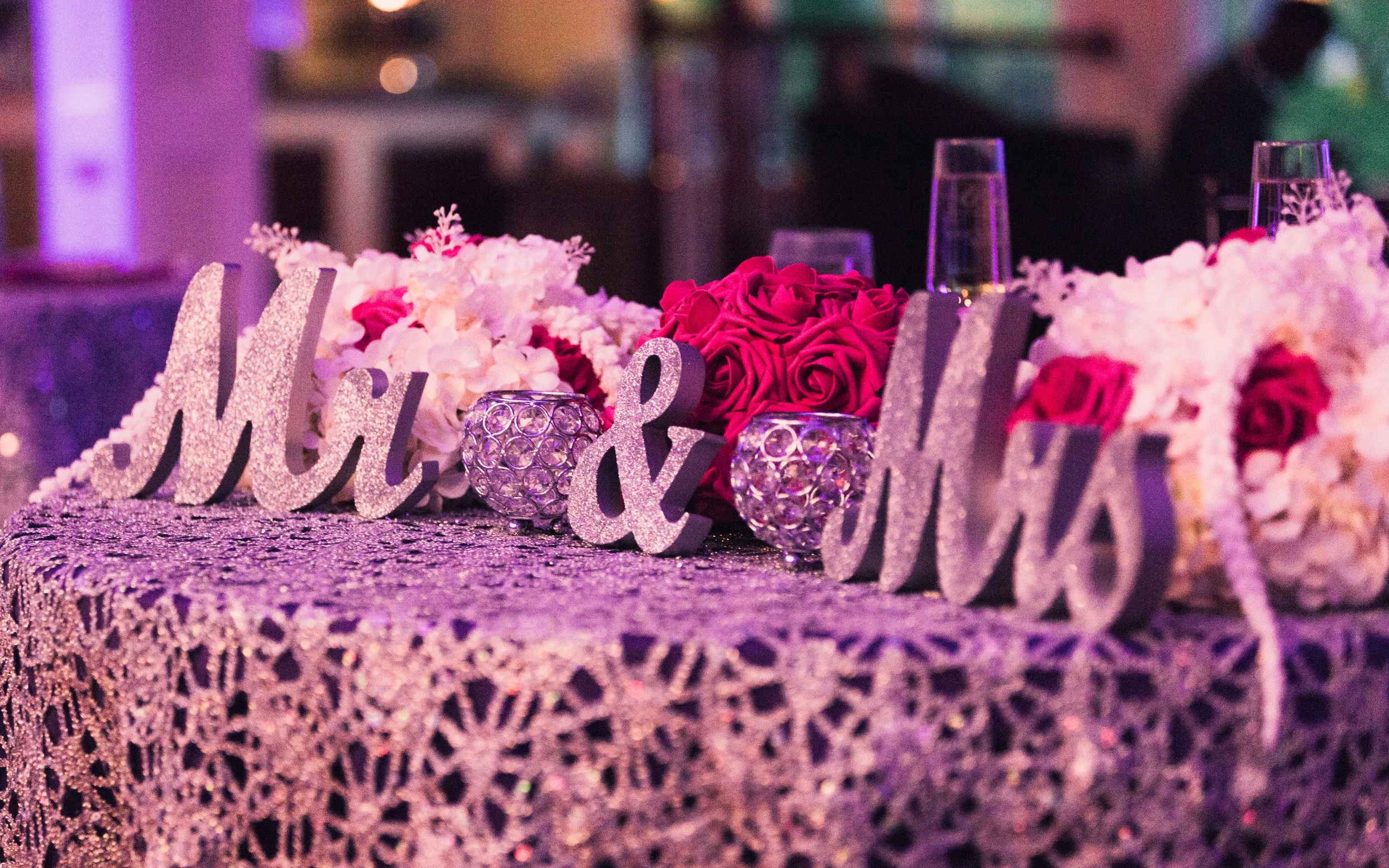 Table decorated with pink roses, white flowers, and glittery 'Love' sign, with glasses and decorative candles, at an event with purple lighting.