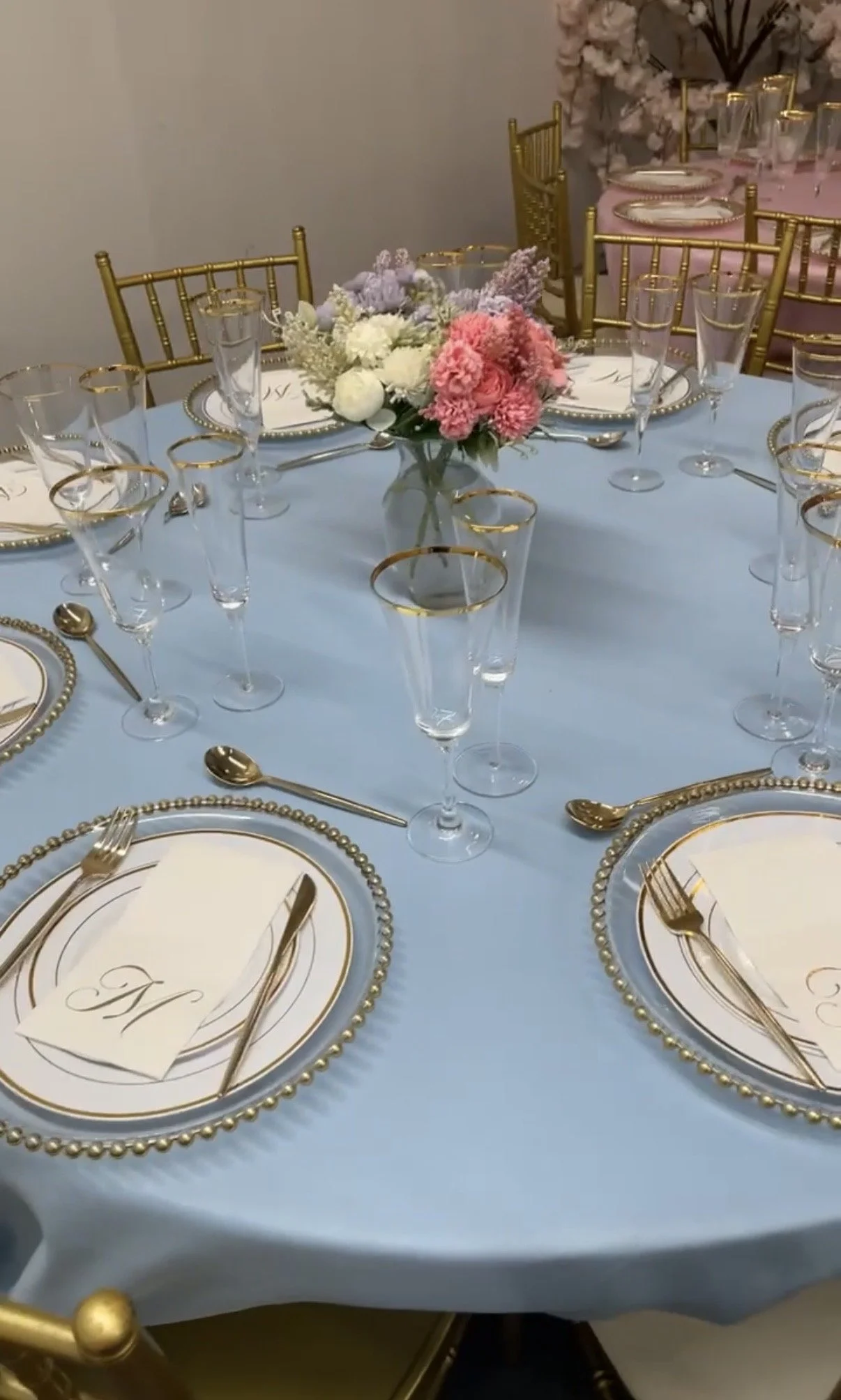 A formal banquet table with a light blue tablecloth, gold and white dinnerware, gold flatware, and clear glasses with gold rims. The place settings include a menu card with a gold initial. In the center of the table is a floral arrangement with pink, purple, and white flowers. Gold chairs surround the table, and another similarly decorated table is visible in the background.