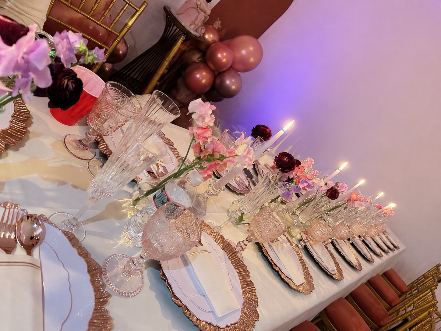 Elegant banquet table decorated with pink and purple flowers, gold-trimmed plates, crystal glasses, candles, and balloons in the background
