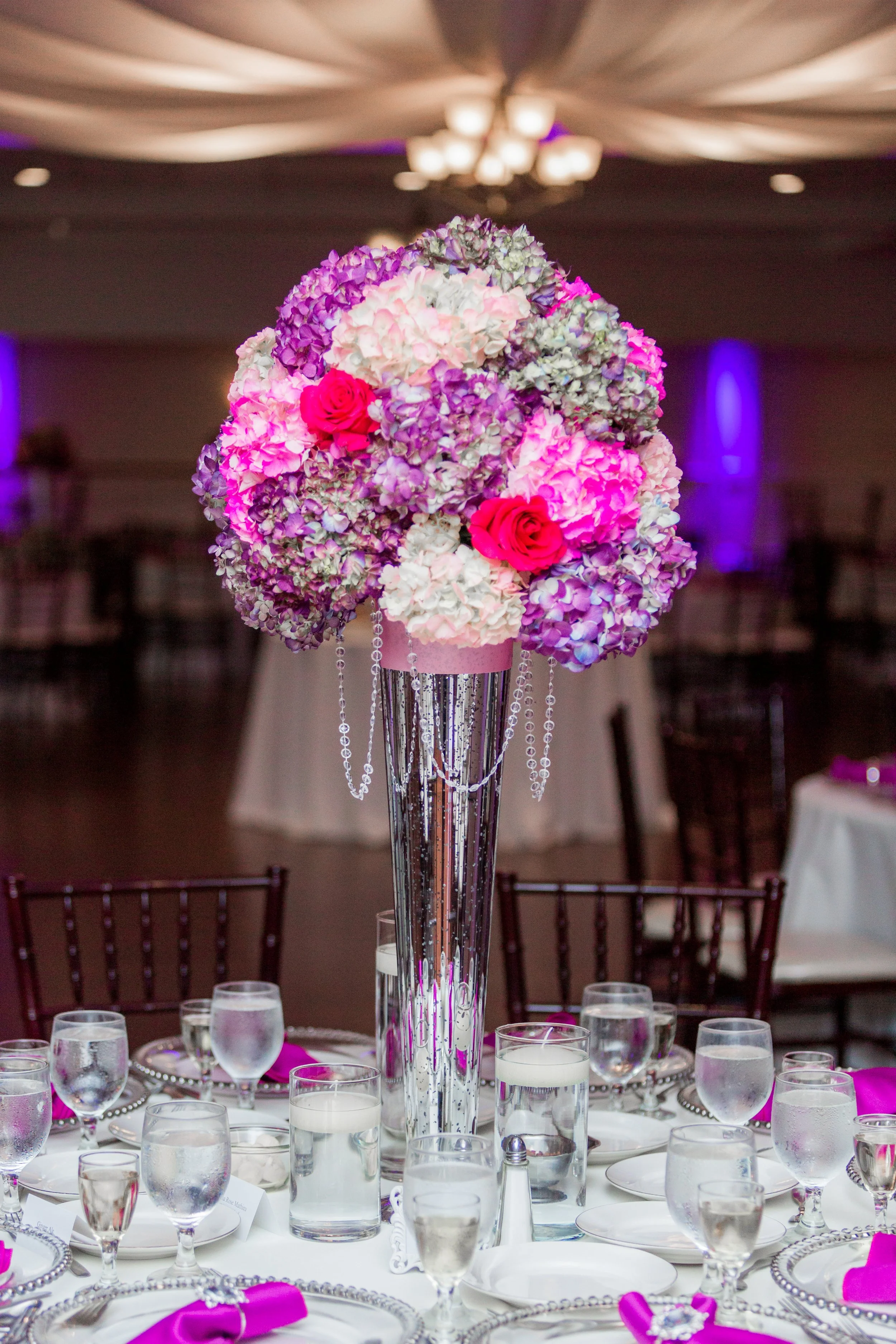 A tall floral centerpiece with pink, purple, white hydrangeas, and red roses on a banquet table, with glasses of water, white plates, pink napkins, and silver charger plates, in an event hall with a decorated ceiling and purple lighting.