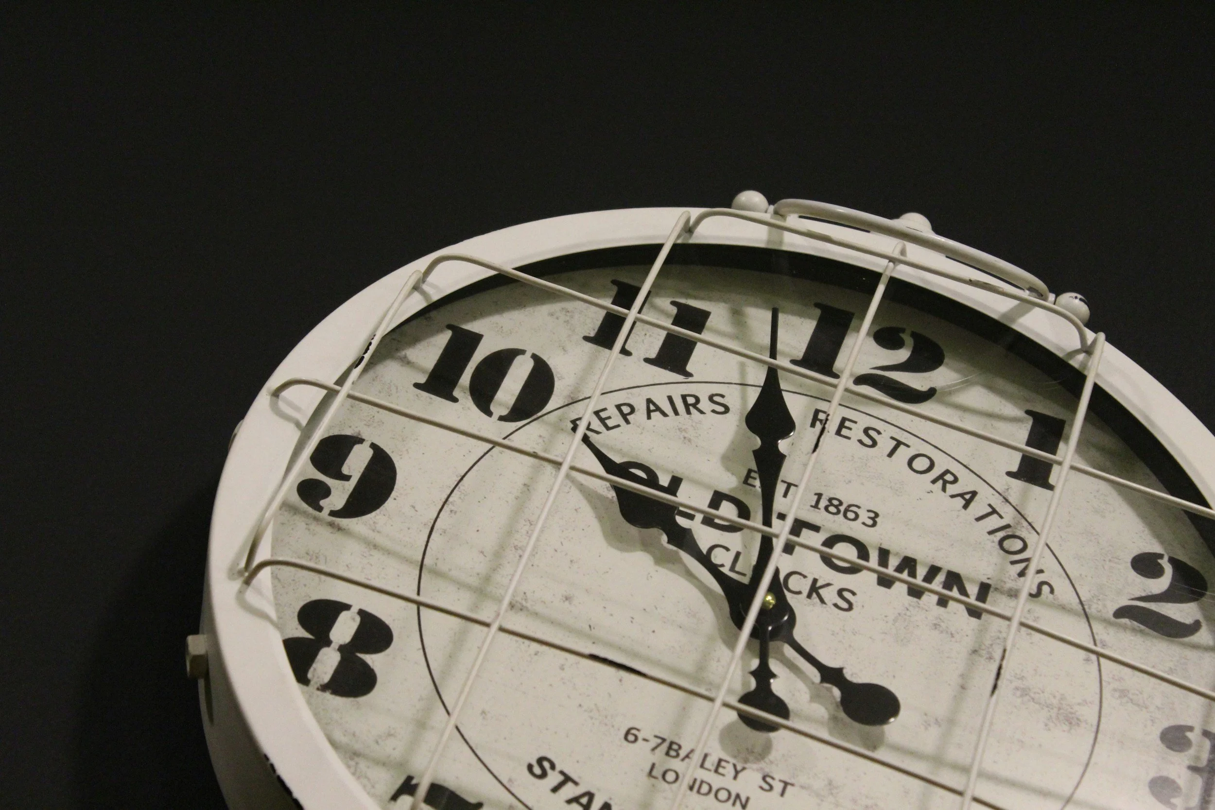 Close-up of a vintage-style wall clock with black numbers on a white face, black hands, and a protective metal grill, against a dark background.