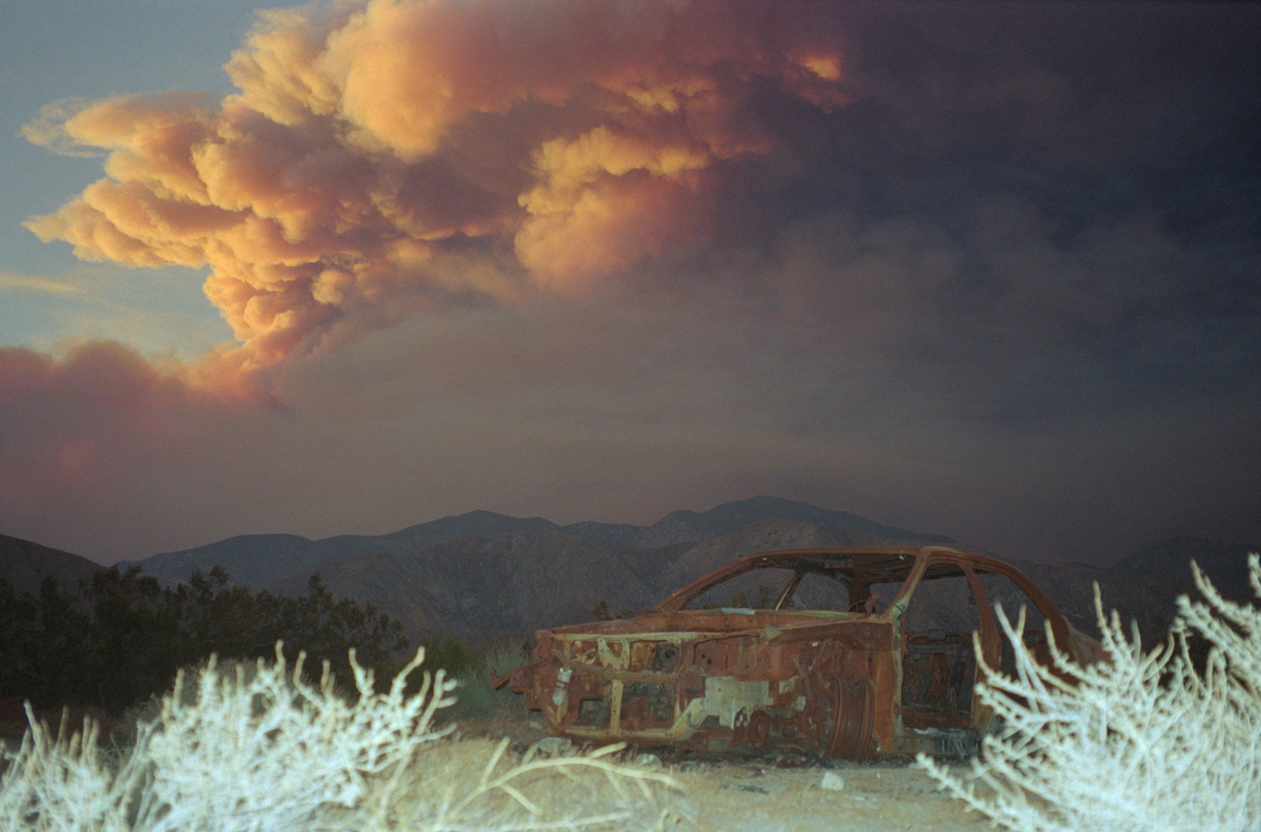 An abandoned, rusted car frame in the foreground amidst desert plants, with mountains in the background and a large, colorful, billowing cloud in the sky.