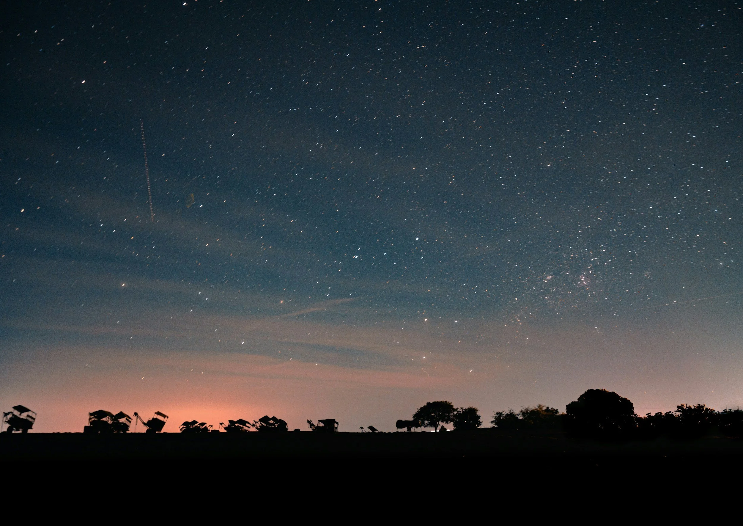 Night sky filled with stars above a horizon with trees and satellite dishes.